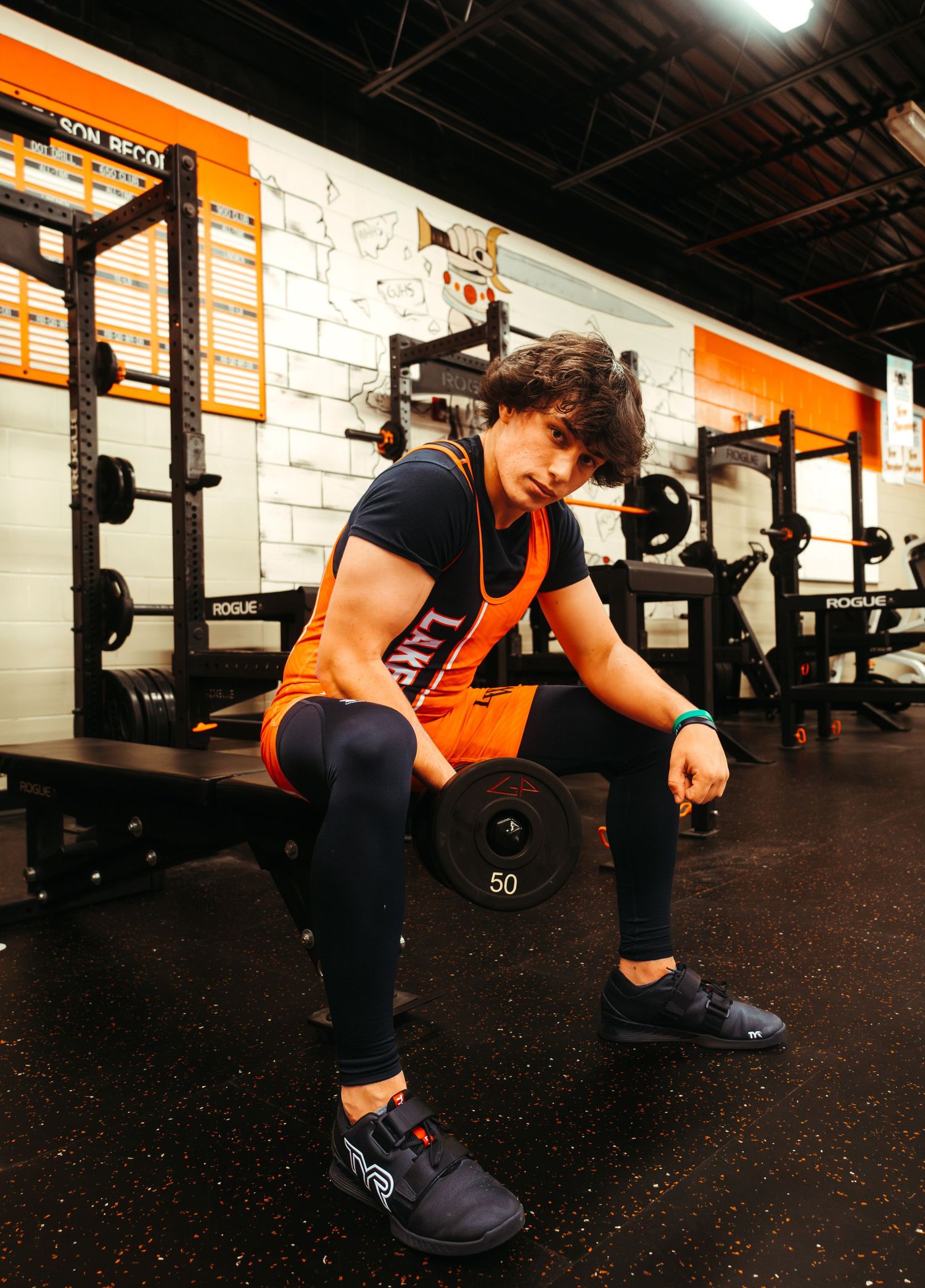 Athlete in orange and black uniform rests on bench between sets at weight training facility.