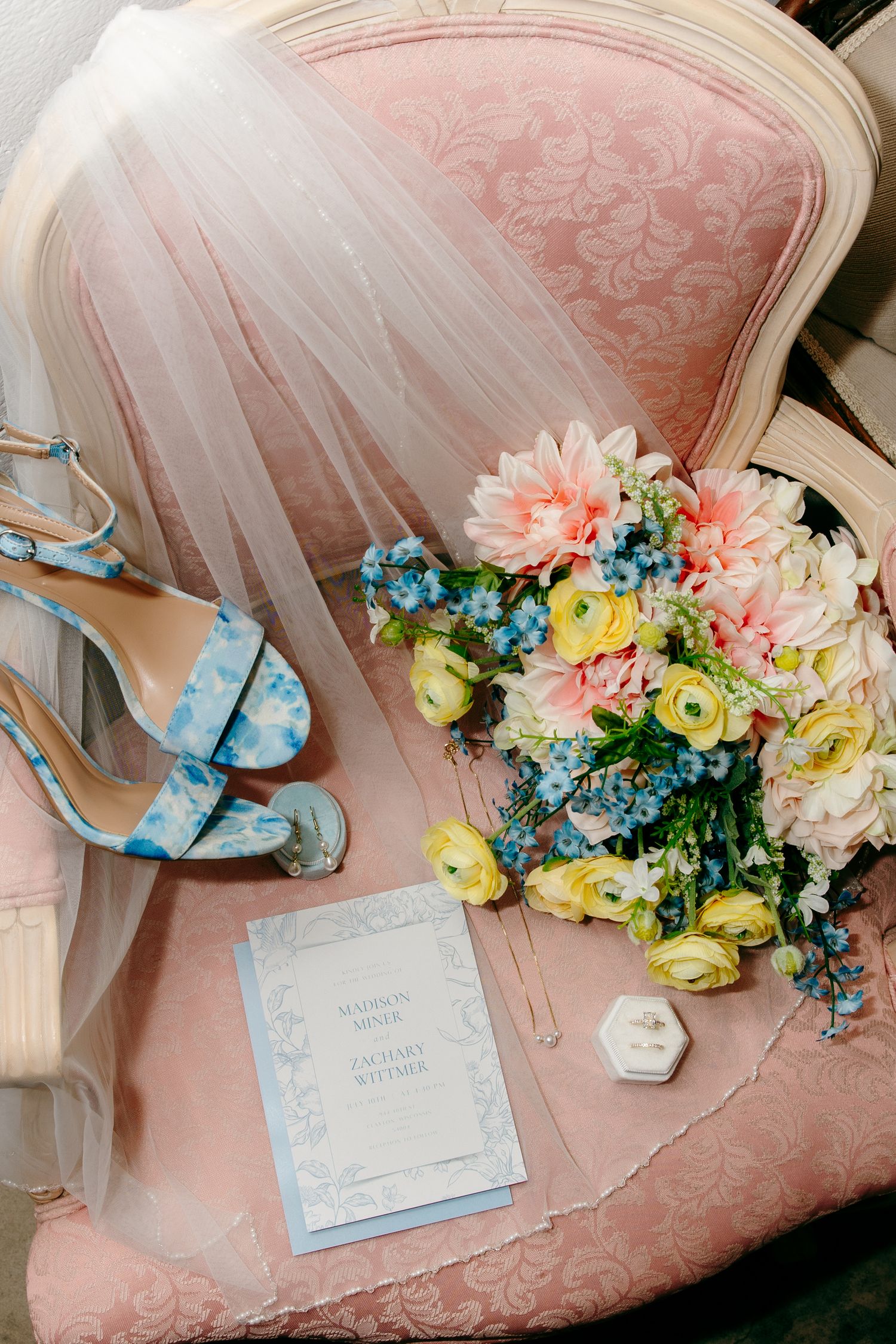 Blue floral heels, pastel bouquet, wedding invitation, and veil arranged on a pink vintage chair.