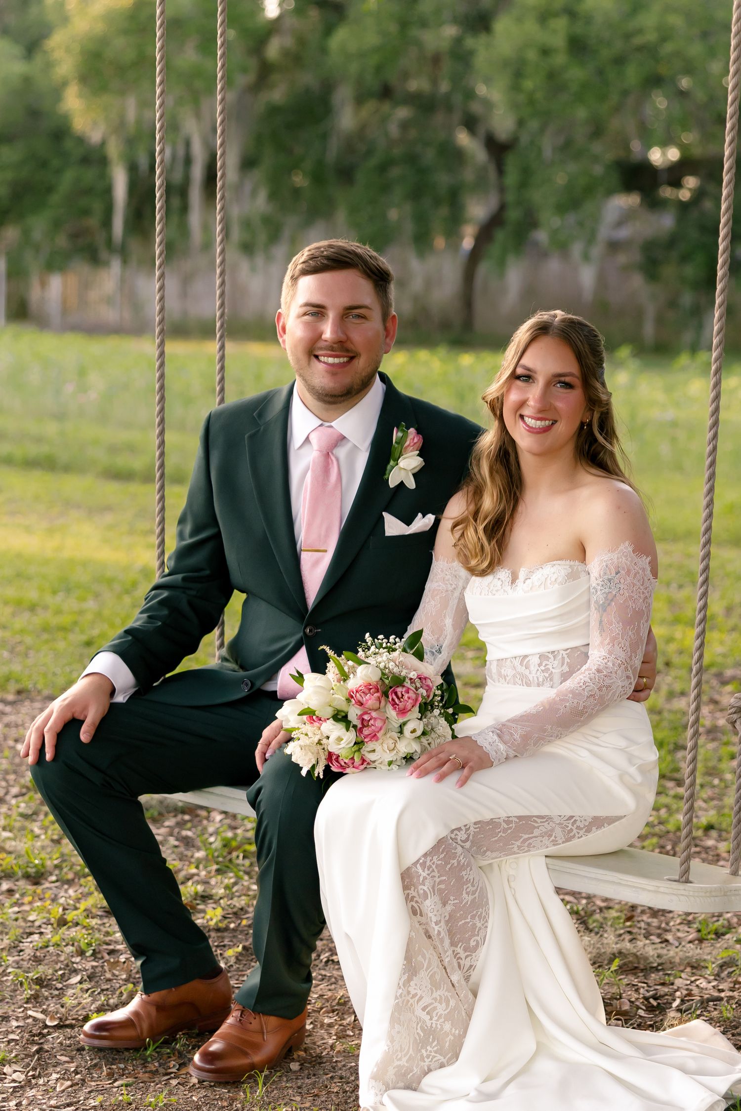 Bride and groom sitting on a swing outdoors, groom in green suit with pink tie, bride in off-shoulder lace gown holding bouquet.