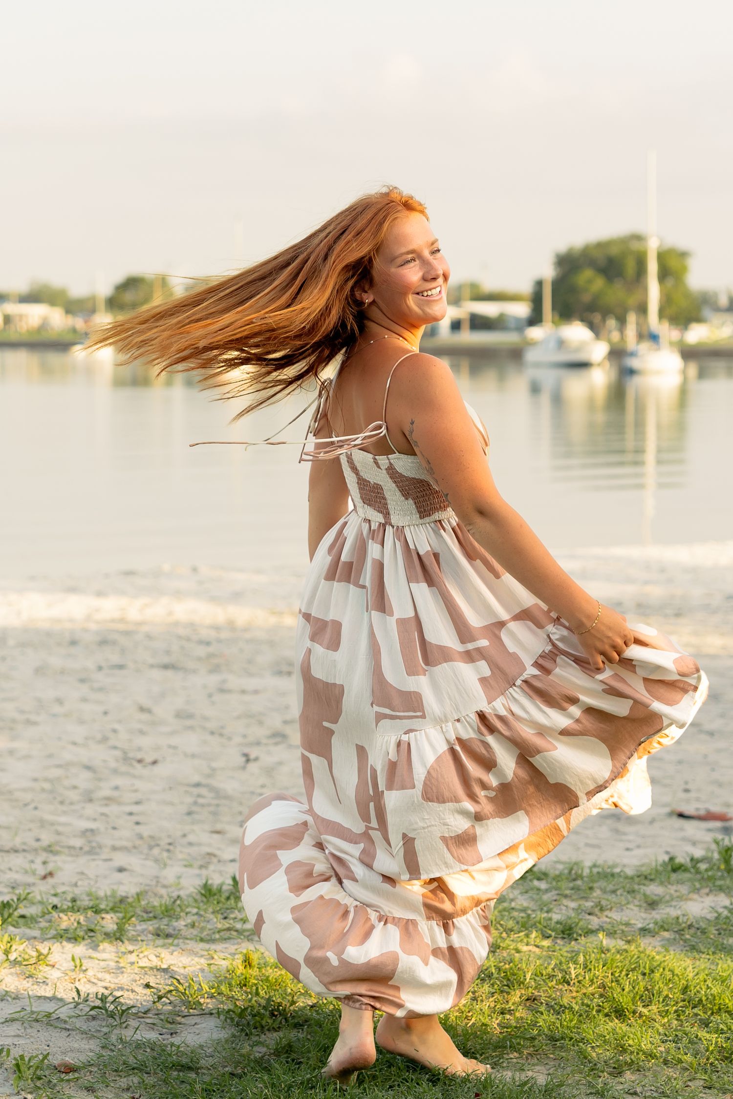 A joyful redhead woman twirls in a flowy boho maxi dress on a grassy beach at golden hour.