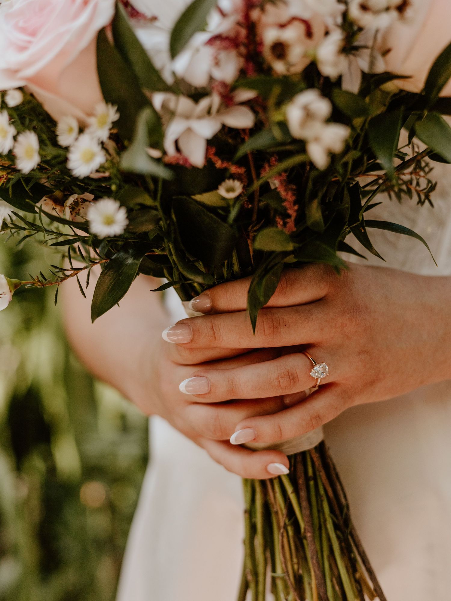 Close up of hands with engagement ring holding a romantic bouquet of pink and white flowers with greenery.