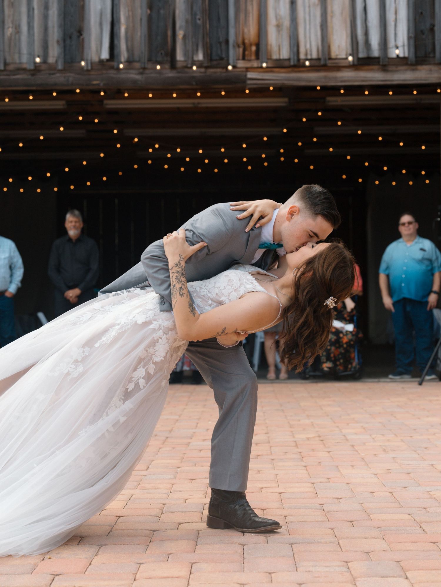 Newlyweds perform romantic dance dip on outdoor brick patio under string lights during their first dance.