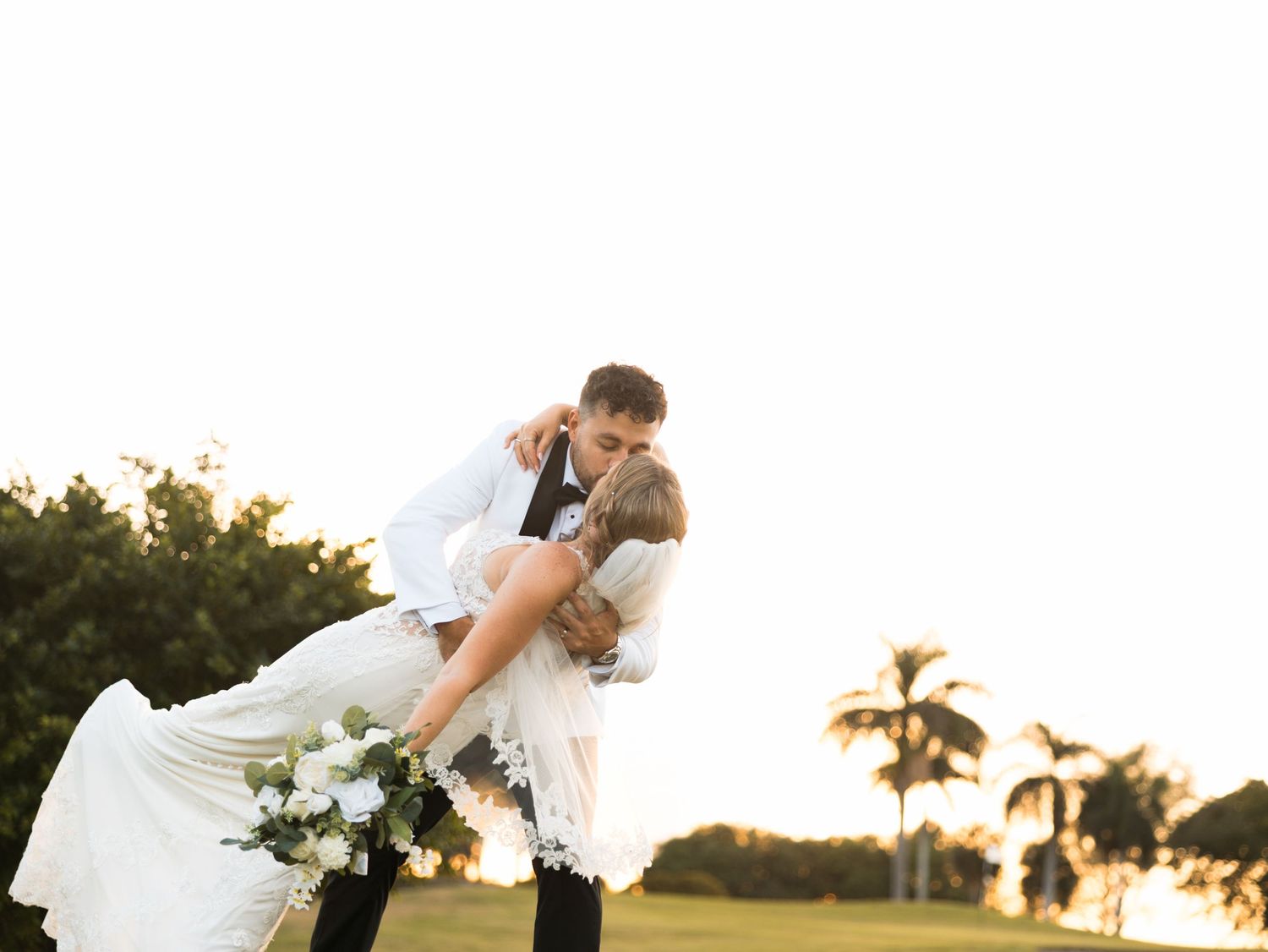 Groom dipping bride in a romantic outdoor wedding kiss, with palm trees and golden sunset in the background.