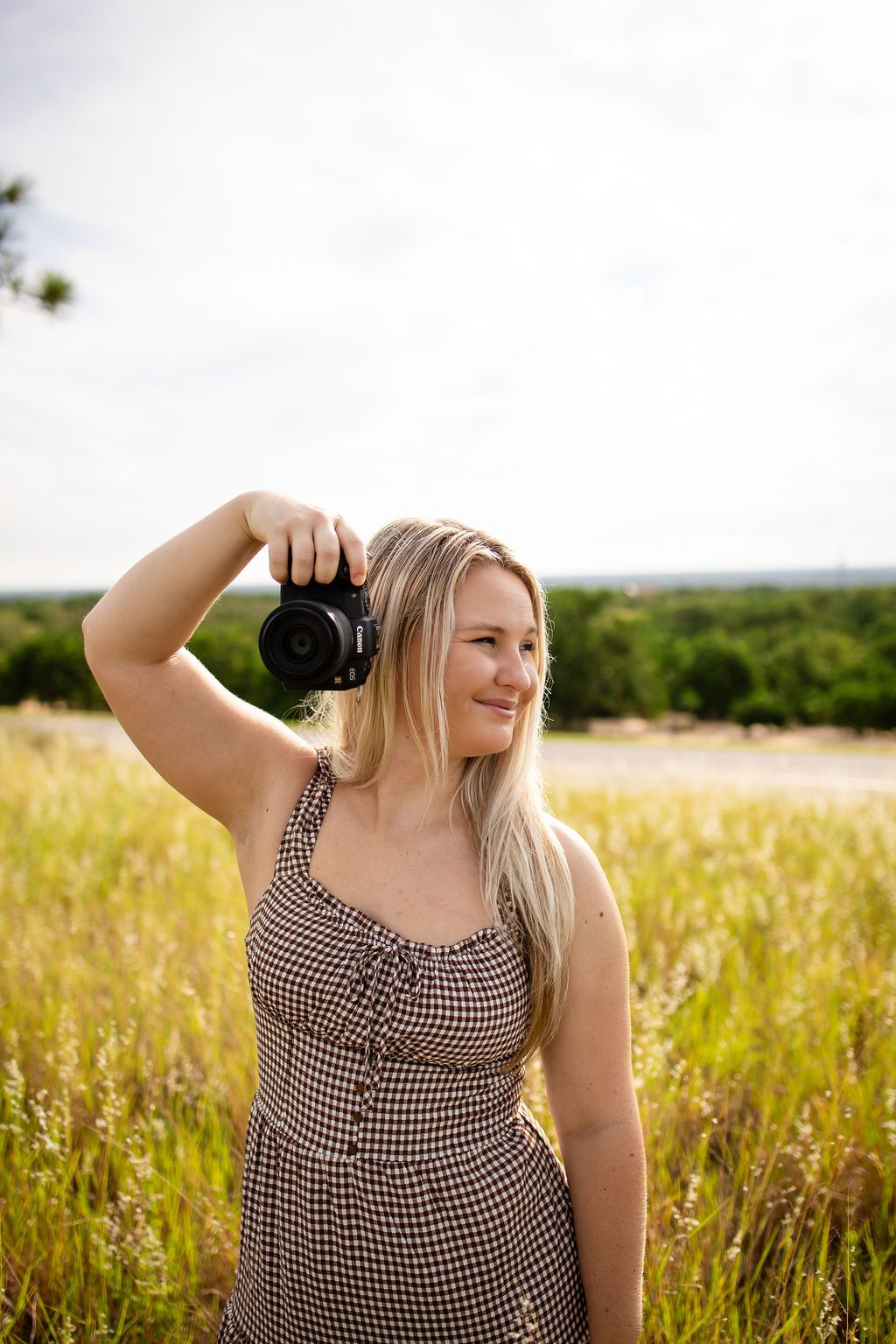 Smiling blonde woman in checkered dress holding DSLR camera in golden field with green trees in background.