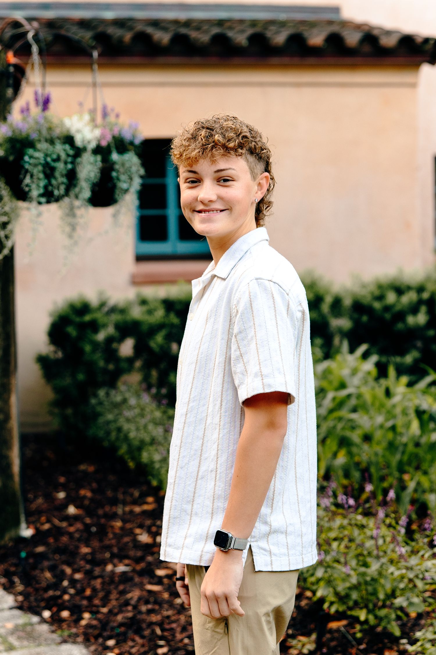Smiling teen with curly hair wearing striped shirt and smartwatch poses outdoors near lush garden and beige building.