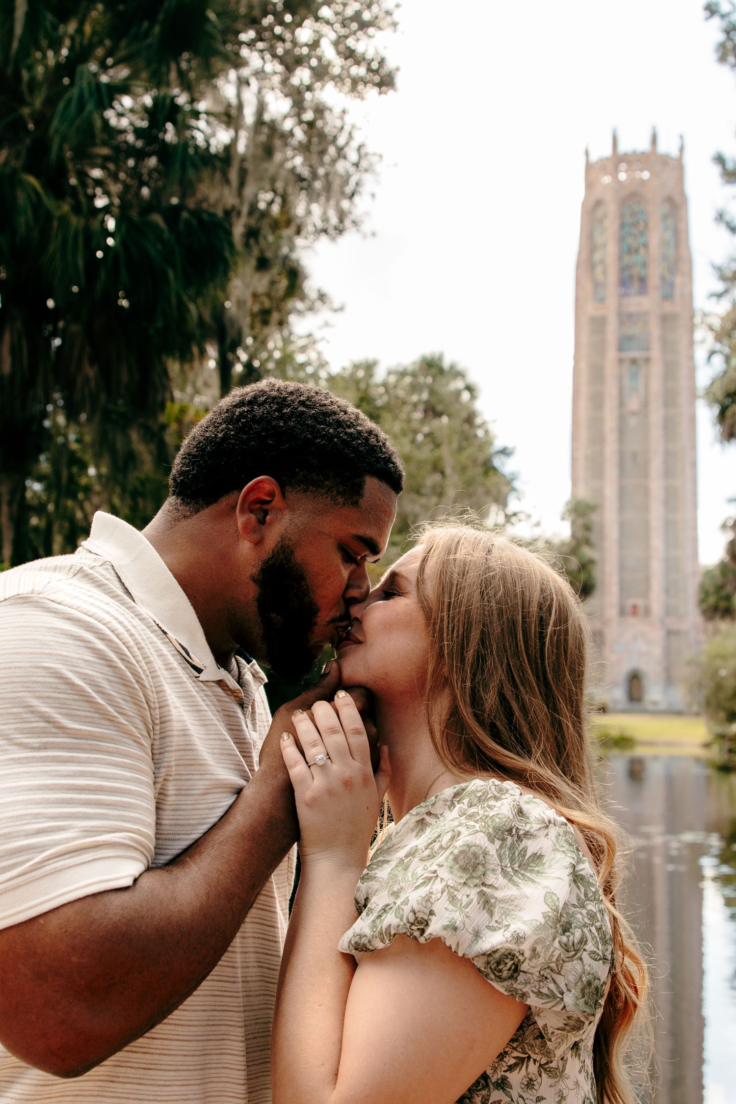 Interracial couple sharing a romantic kiss near a pond with Bok Tower in the background.
