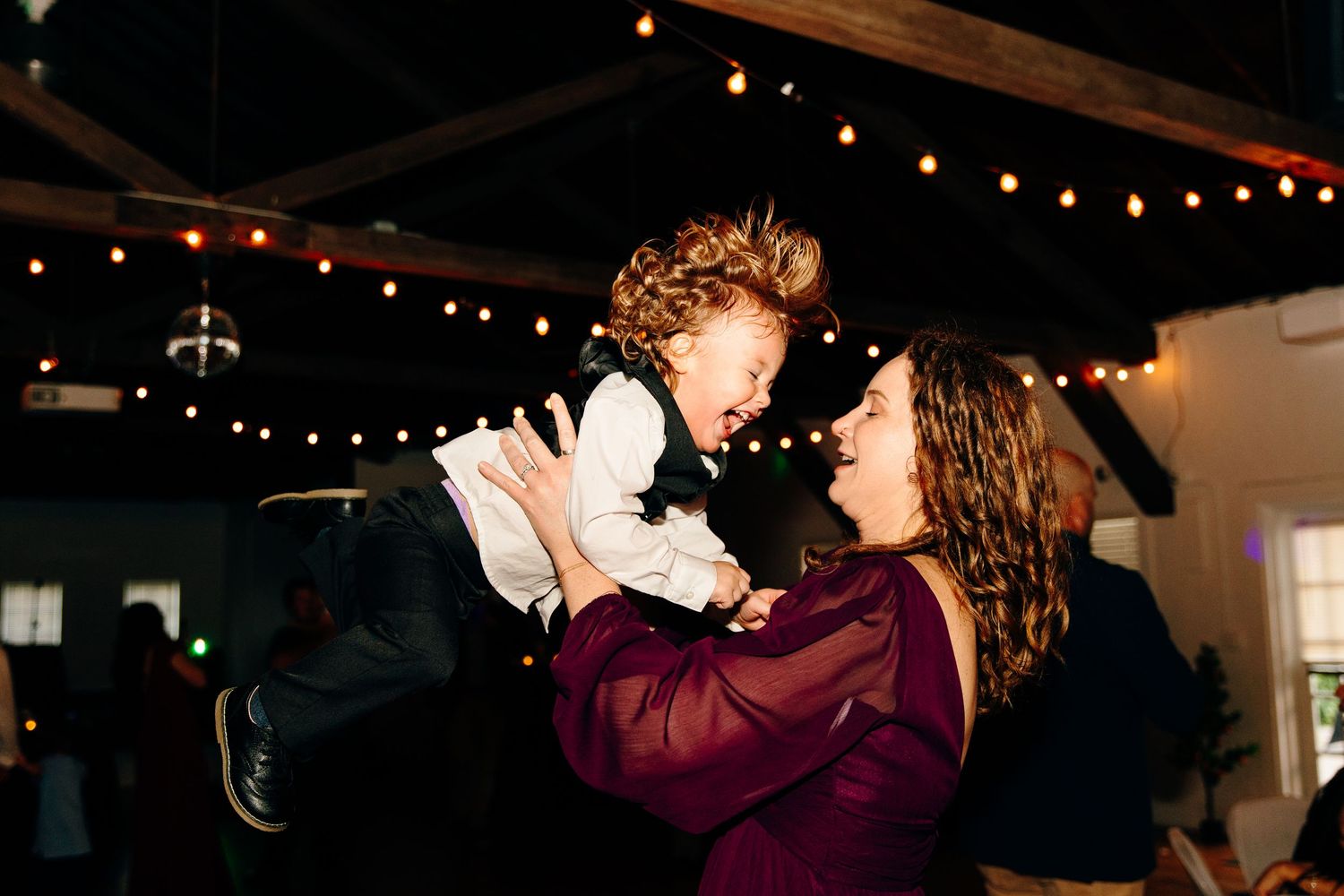 Woman in burgundy dress joyfully lifting laughing child at indoor wedding reception with string lights.