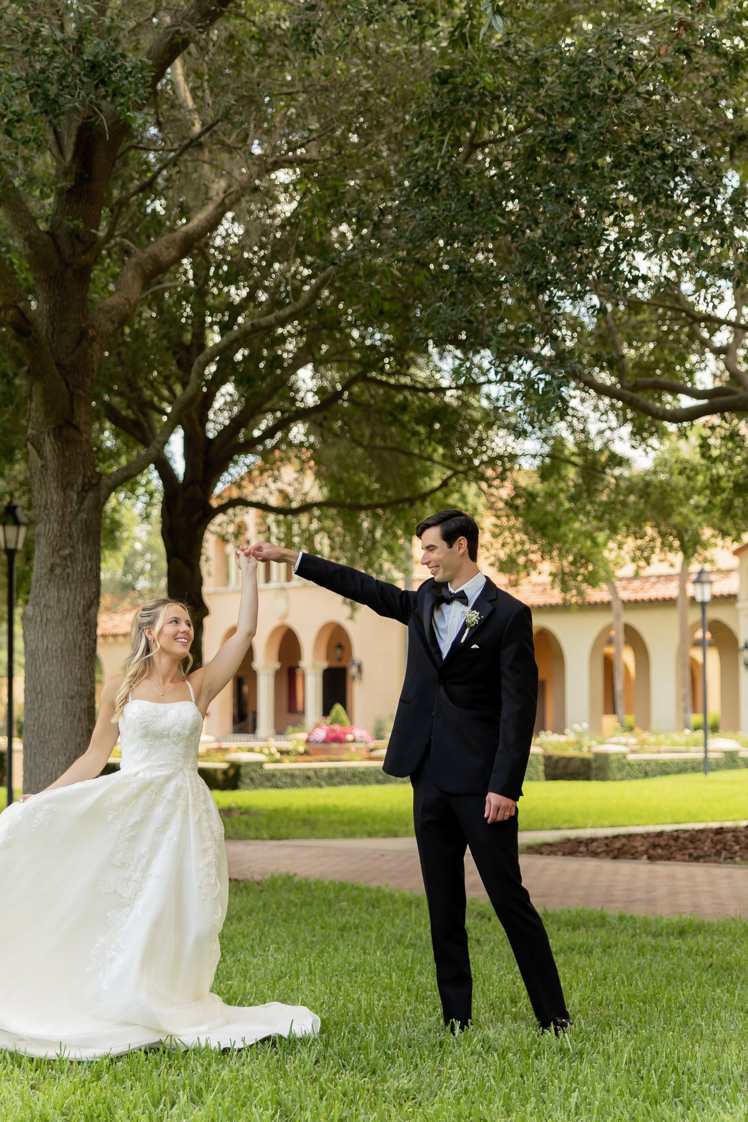 Bride in white gown and groom in black tuxedo dancing on green lawn with historic building backdrop.