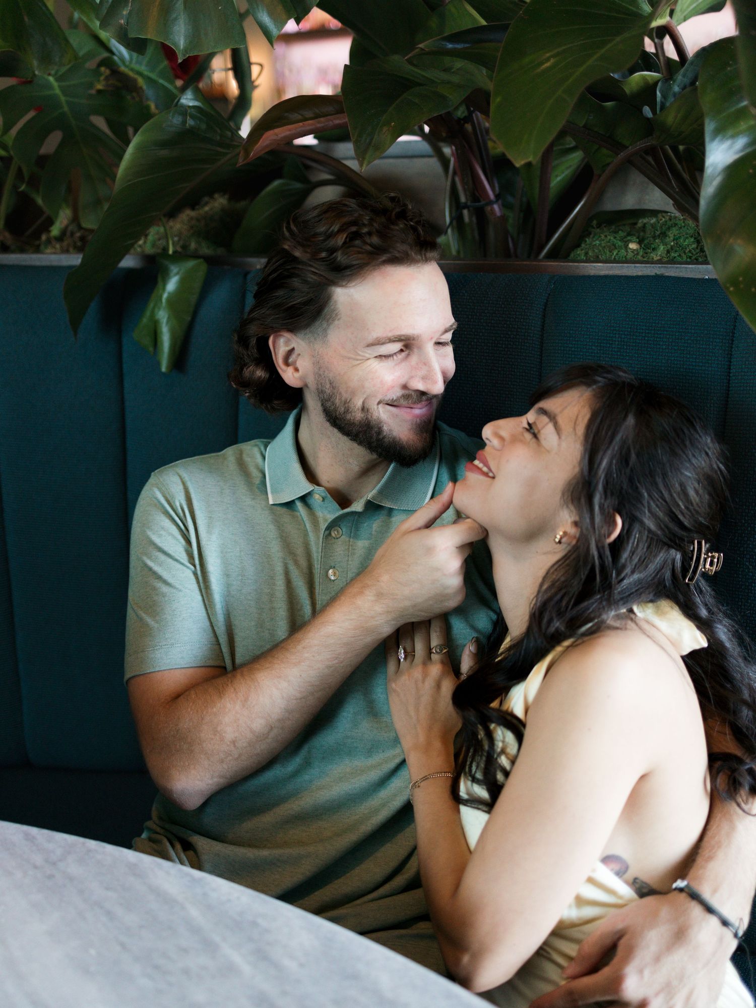 A couple shares an intimate moment while sitting together at a restaurant booth surrounded by green plants.