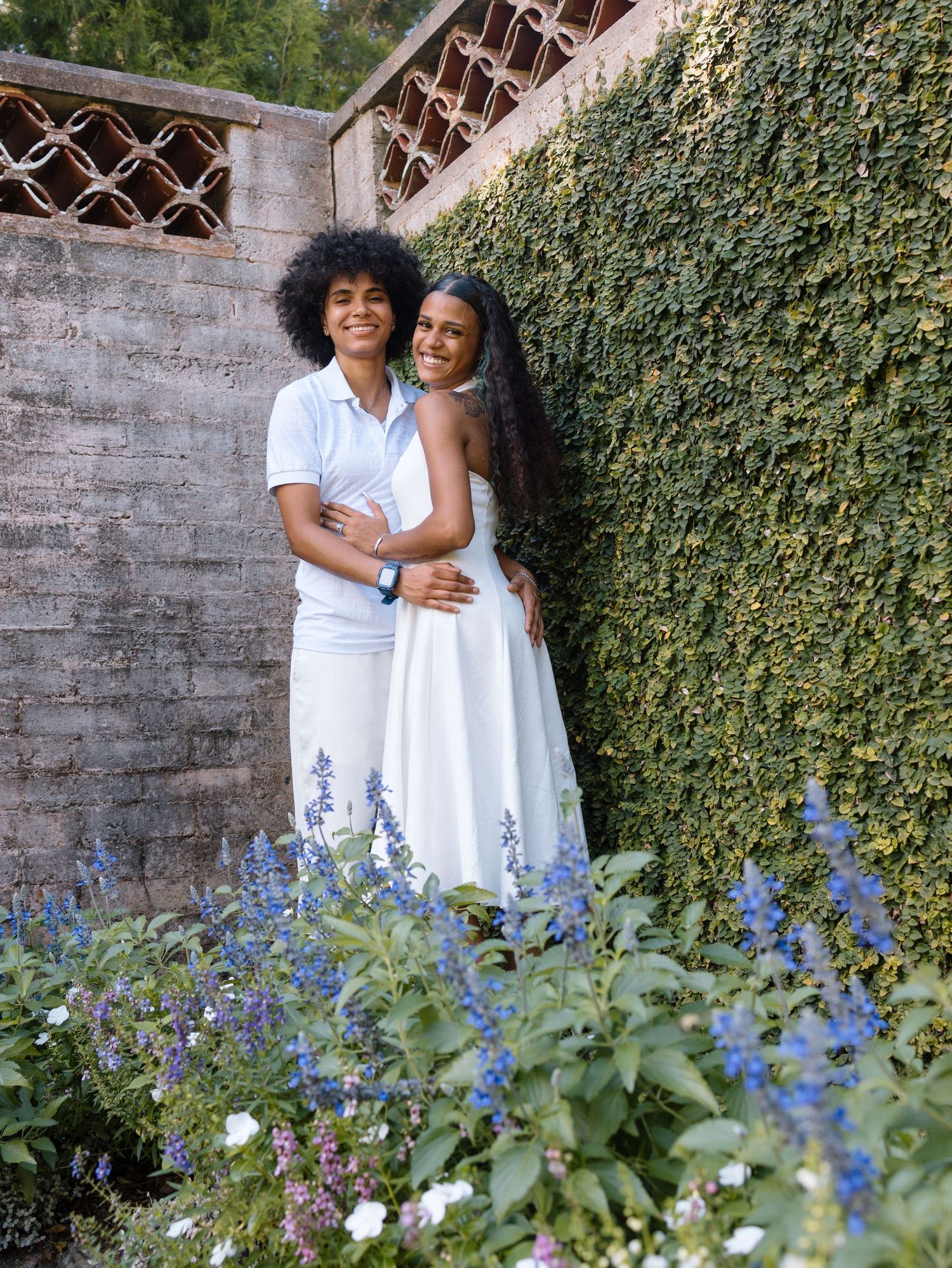 A pair in white sundresses stand together against a brick wall with flowering vines creating a lovely garden backdrop.
