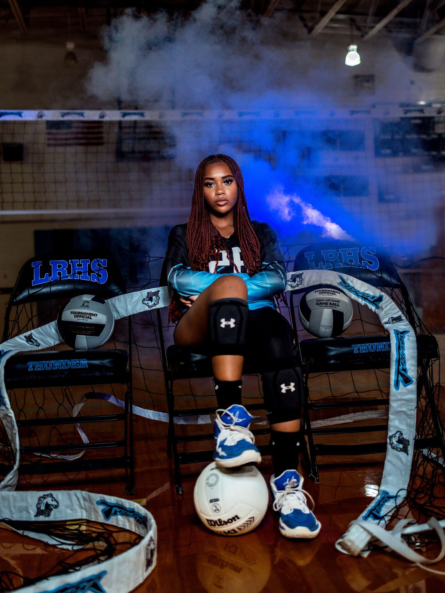 Volleyball player wears black Under Armour gear against dramatic blue-lit background with sports equipment.