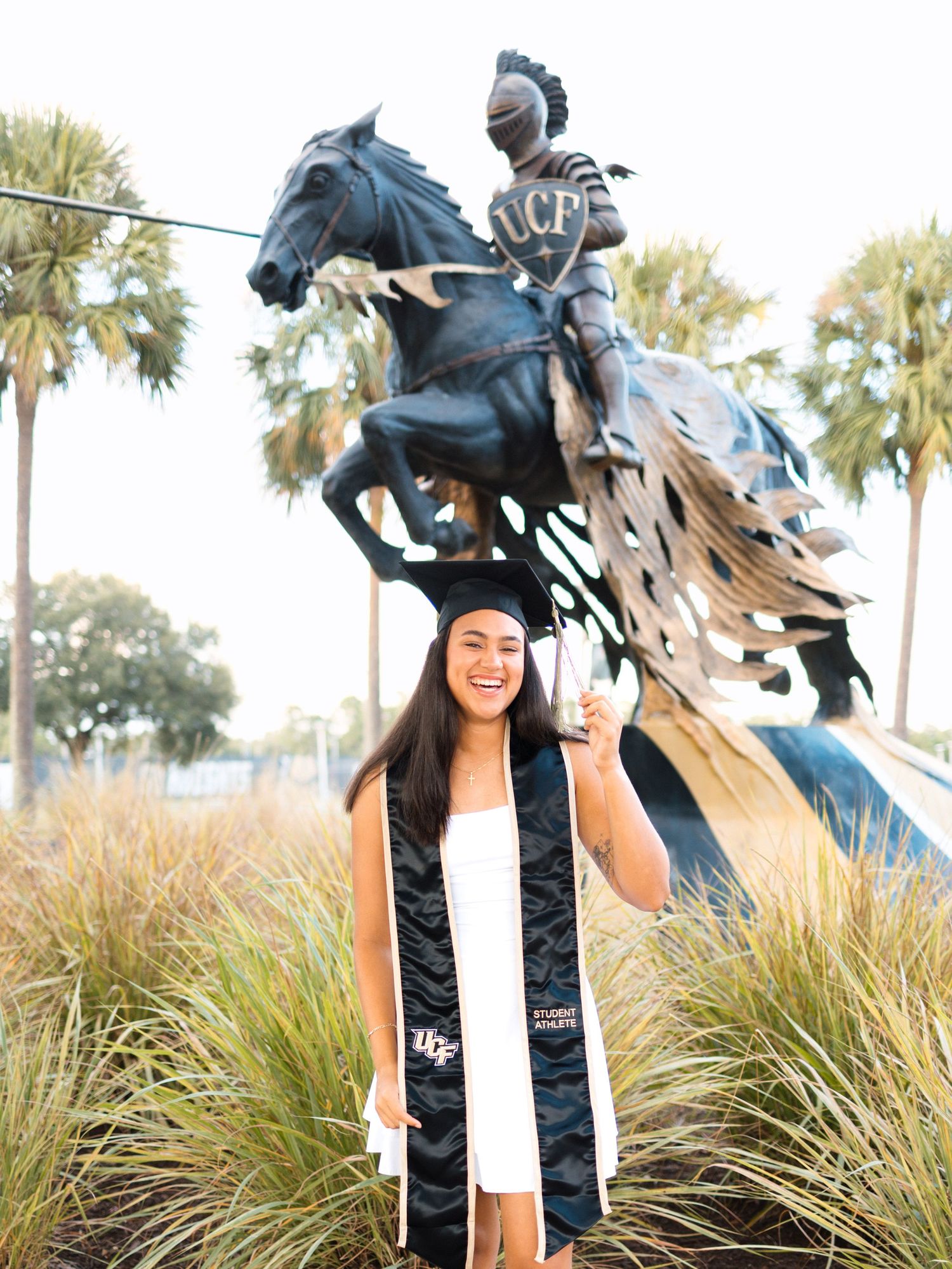 UCF graduate poses proudly in cap and gown stole before the iconic Knight on horseback statue on campus.