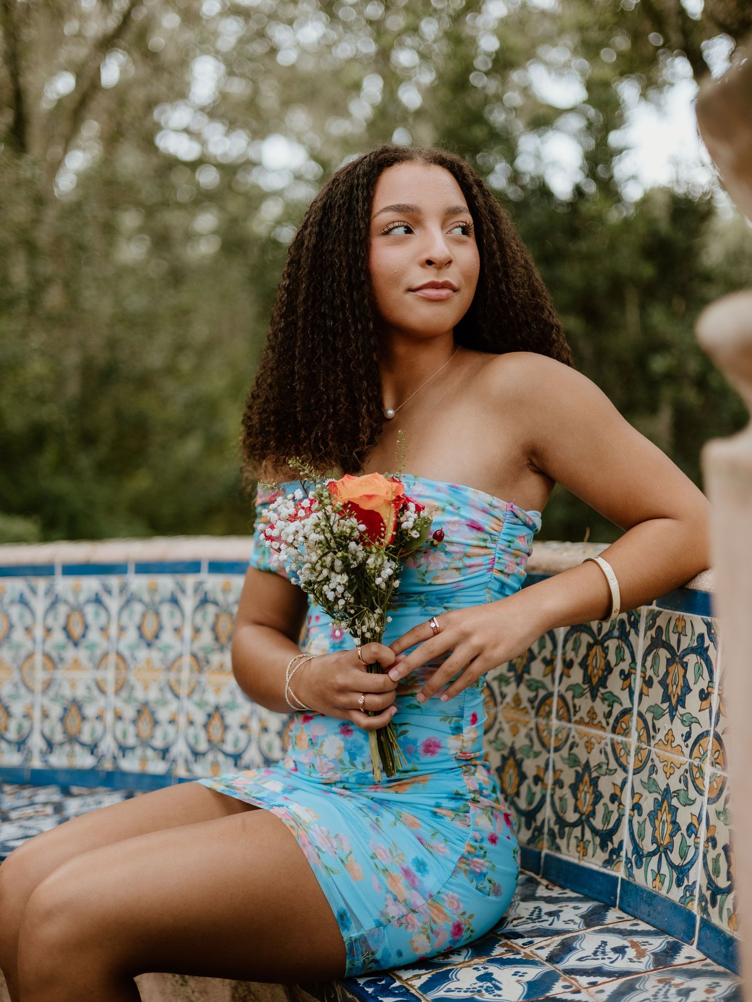 Young woman in floral blue strapless dress holding bouquet, seated by ornate tiled wall outdoors.