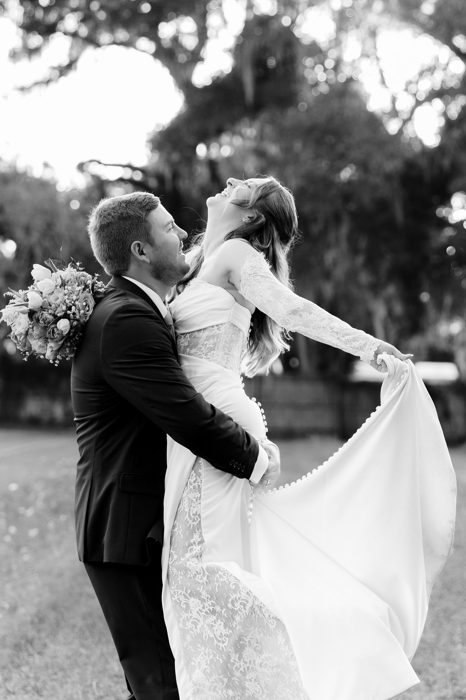 Black and white photo of a groom lifting his bride in a lace wedding dress outdoors under large trees.