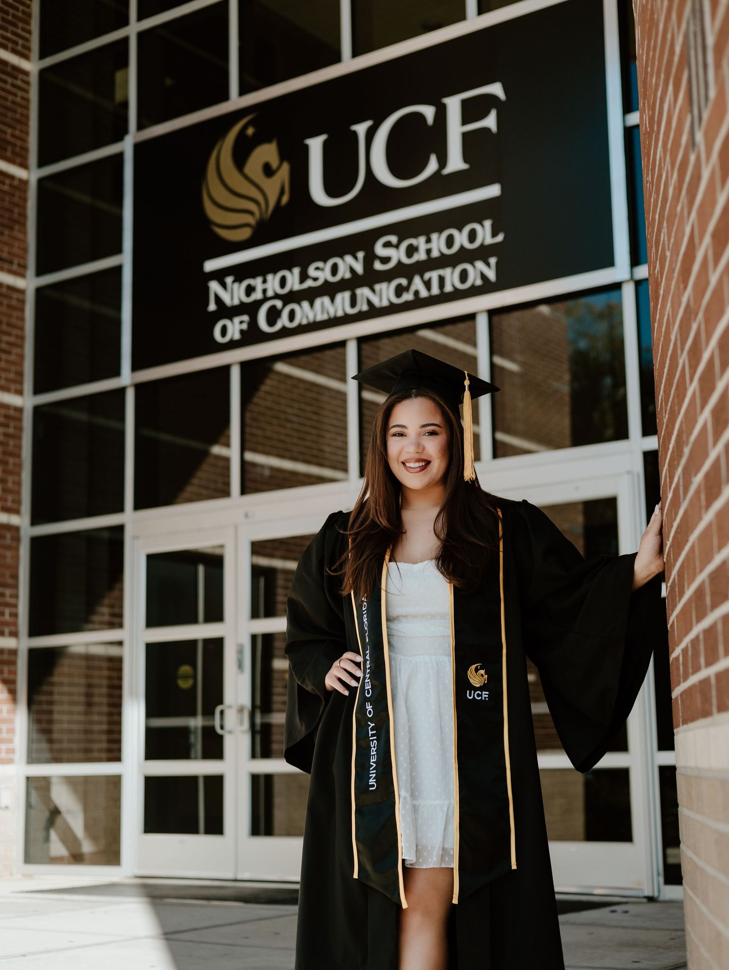 UCF graduate in black cap and gown poses proudly outside Nicholson School of Communication building at University of Central Florida.