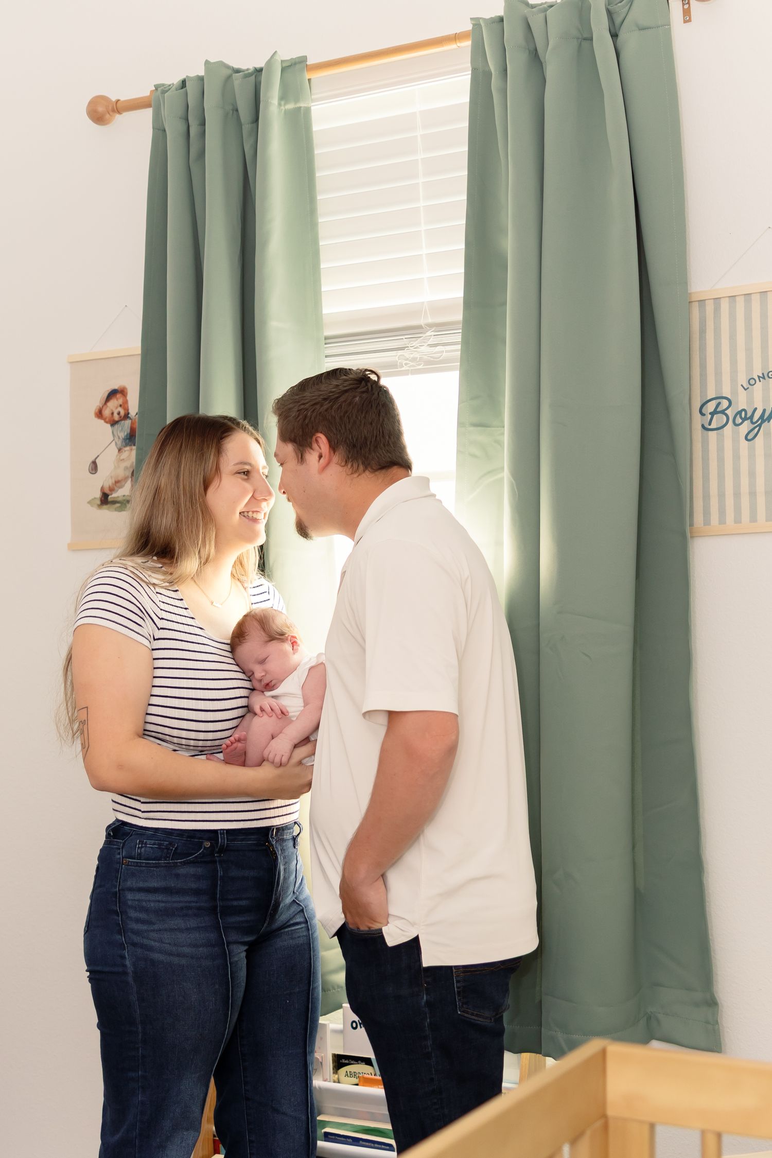 A couple smiles at each other while holding their newborn baby in a nursery with green curtains.