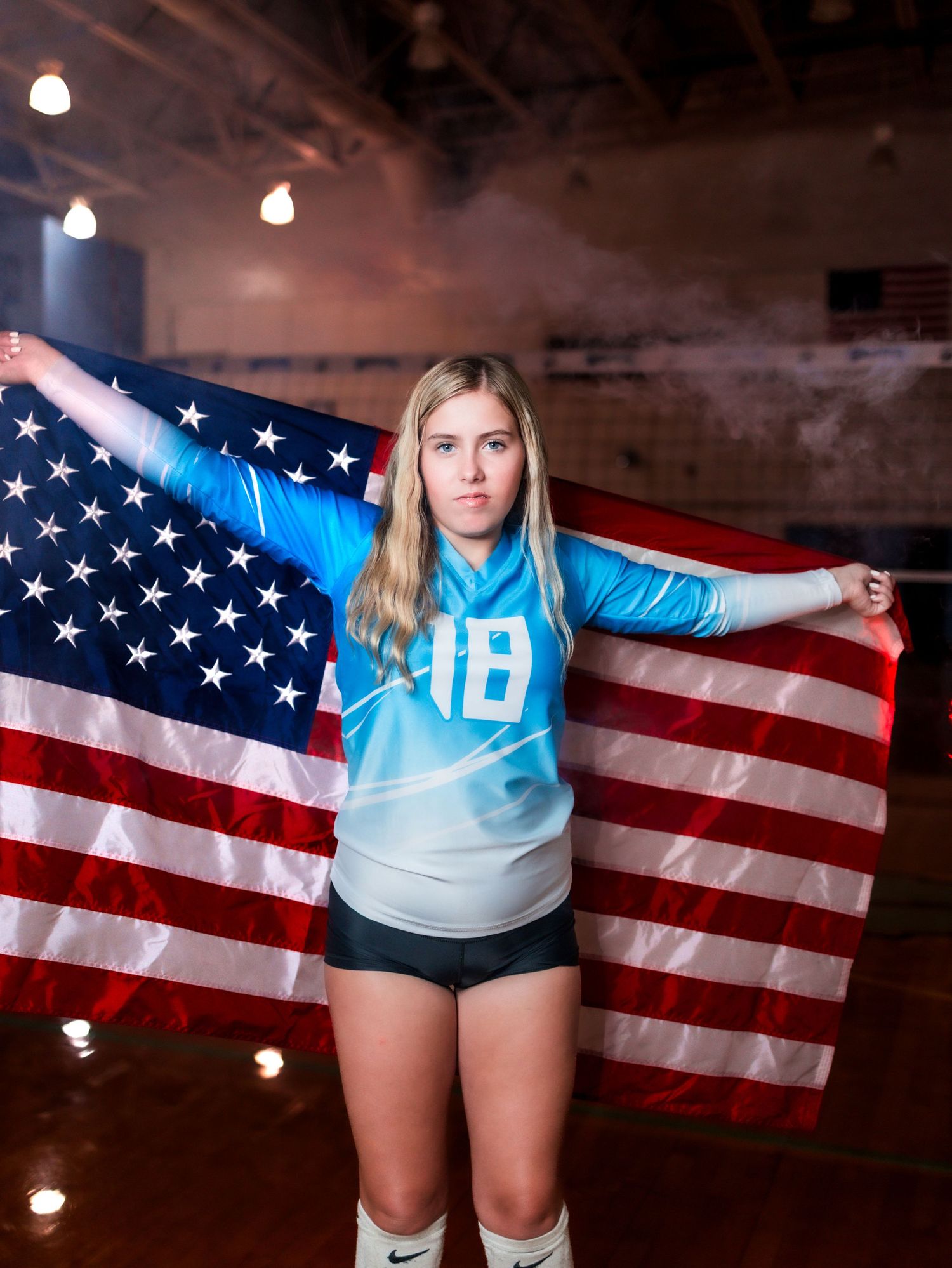 Volleyball player poses with American flag in indoor gym wearing light blue uniform against dark background.