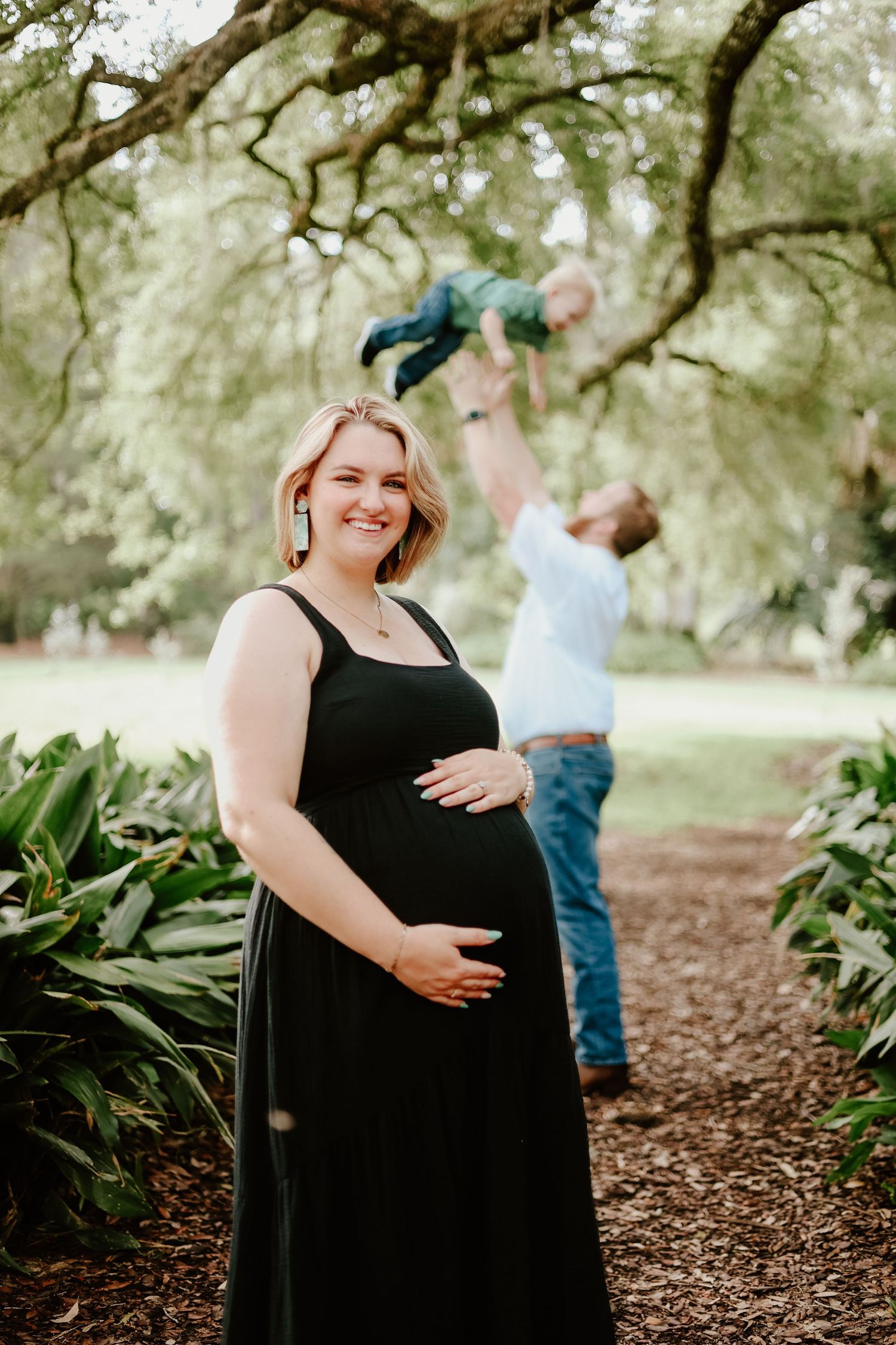 A series of maternity photos showing a woman in a black dress standing on a wooded path surrounded by trees and foliage.