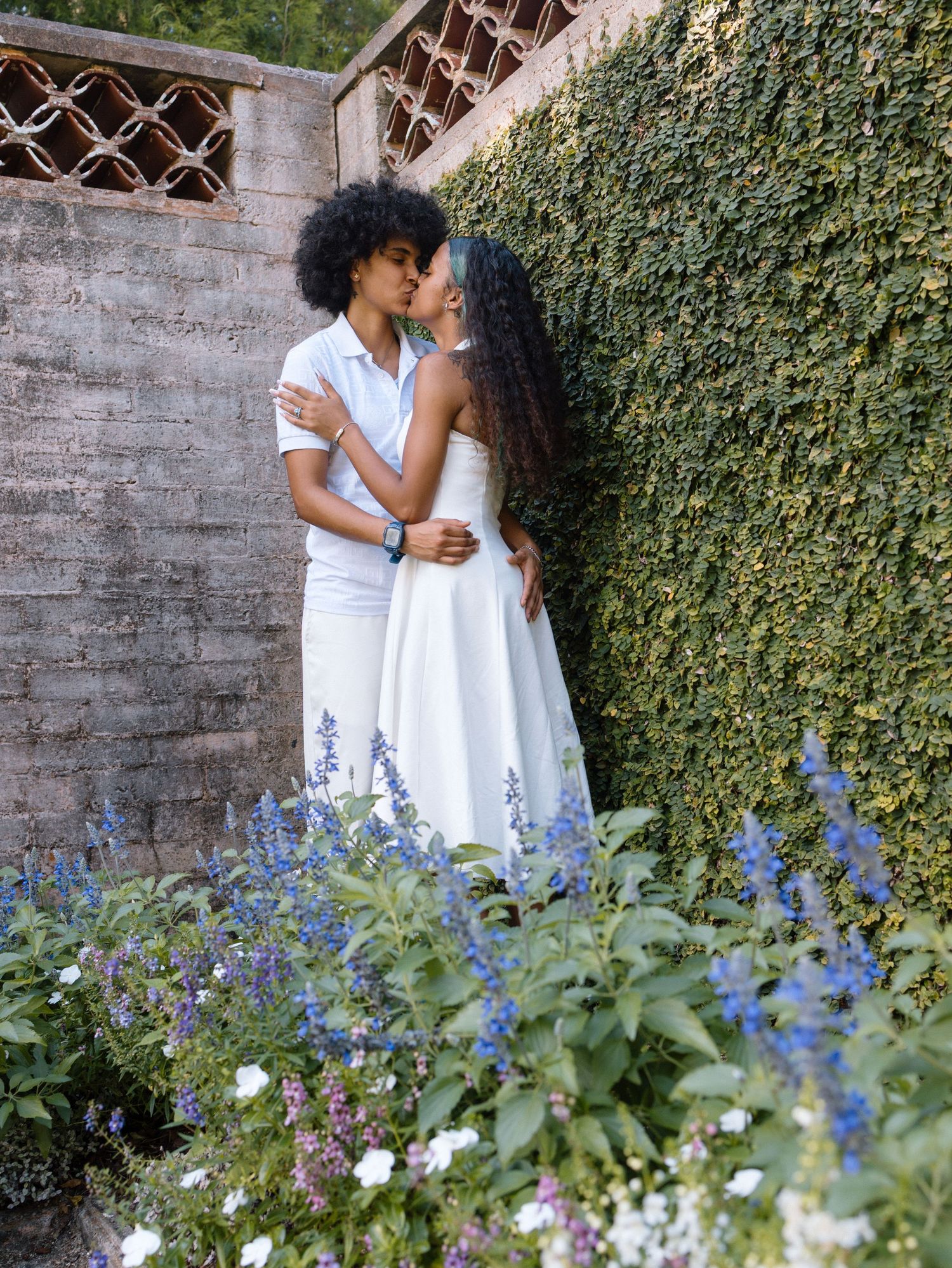 A romantic couple in white outfits embrace near an ivy-covered wall with delicate blue flowers blooming in the foreground.