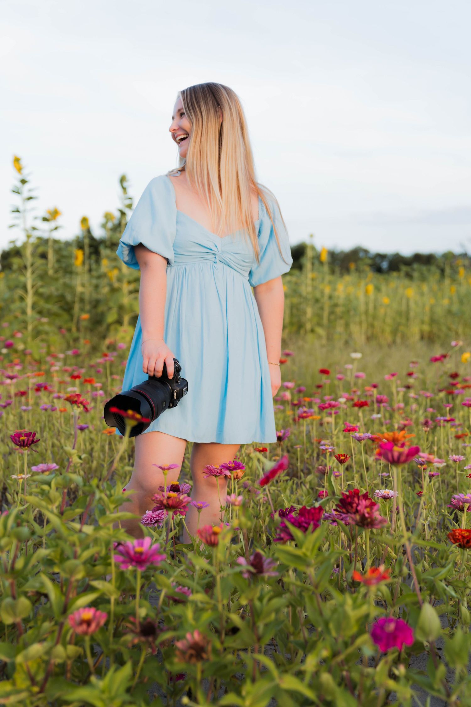 Smiling blonde woman in light blue dress holding a camera standing in a vibrant colorful flower field outdoors.