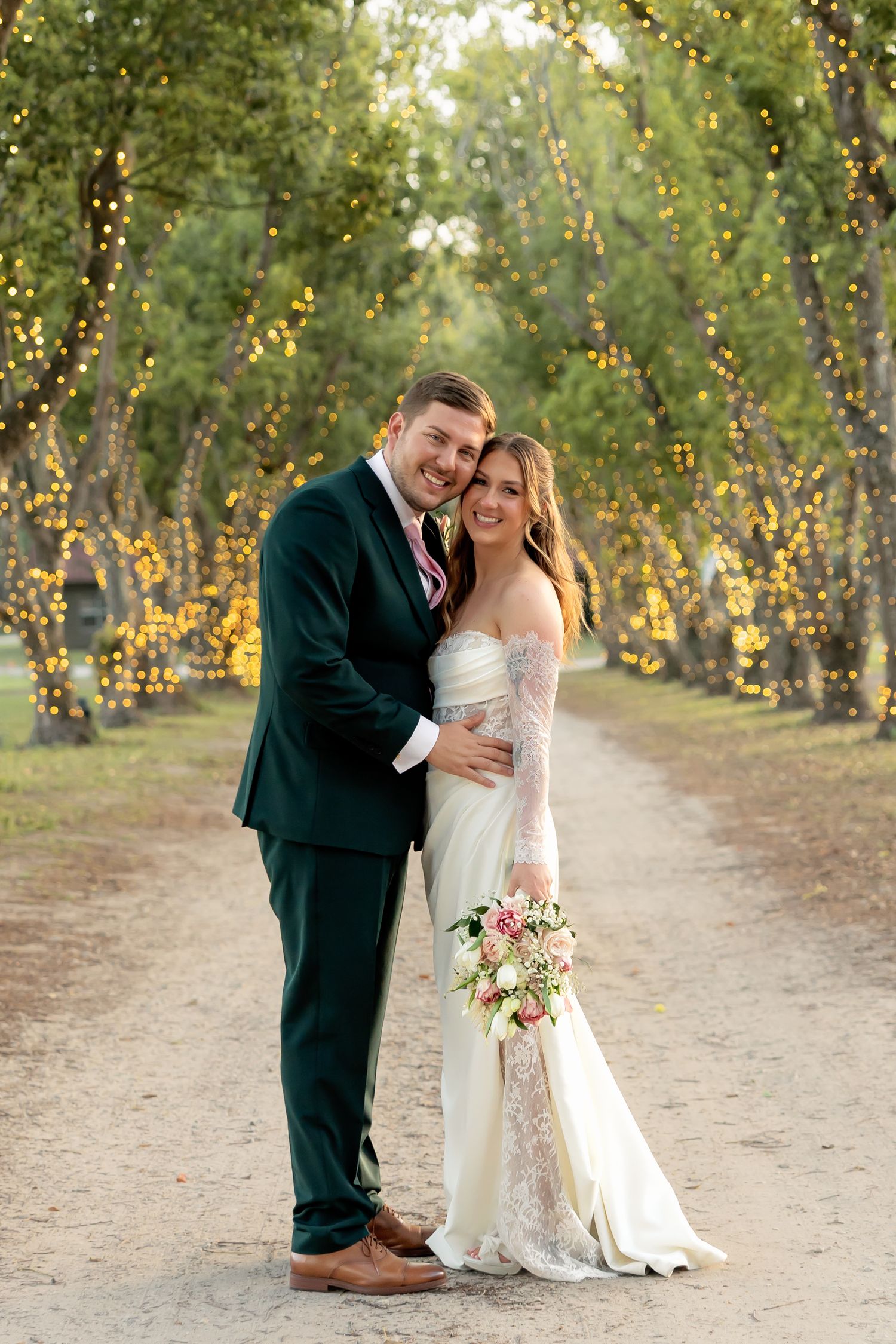 Bride and groom pose on a tree-lined path adorned with warm fairy lights at their outdoor wedding.