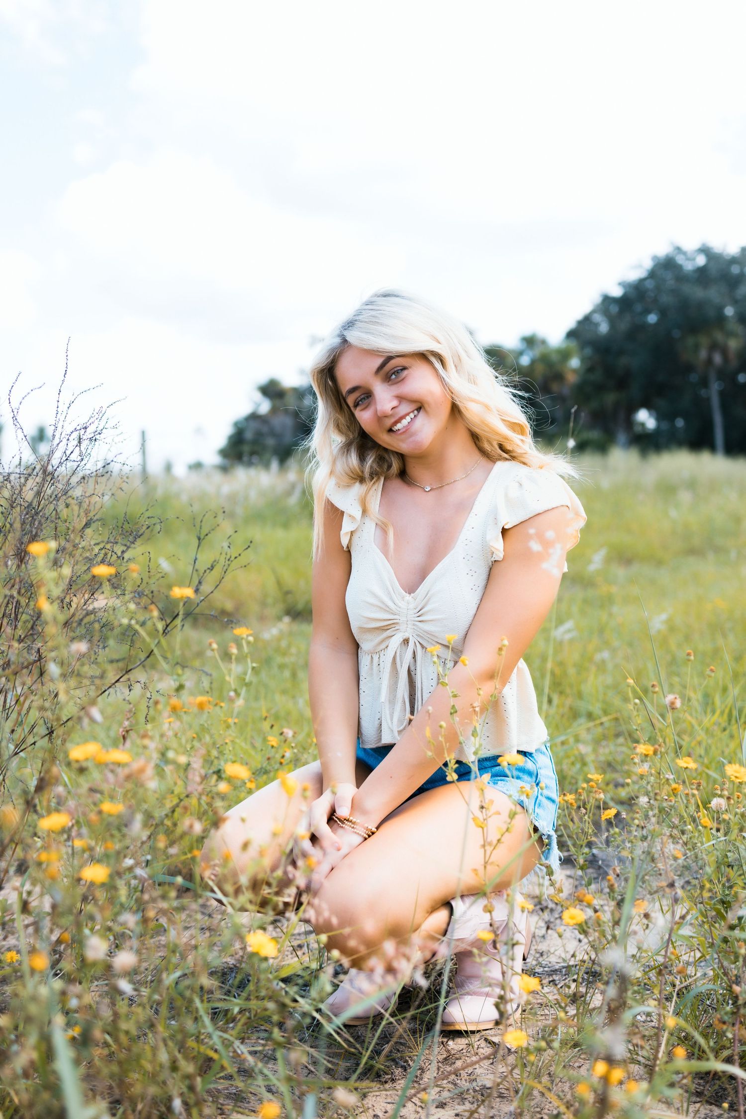 A woman in a white dress sits among wildflowers in a sunny meadow during a summer photo session.