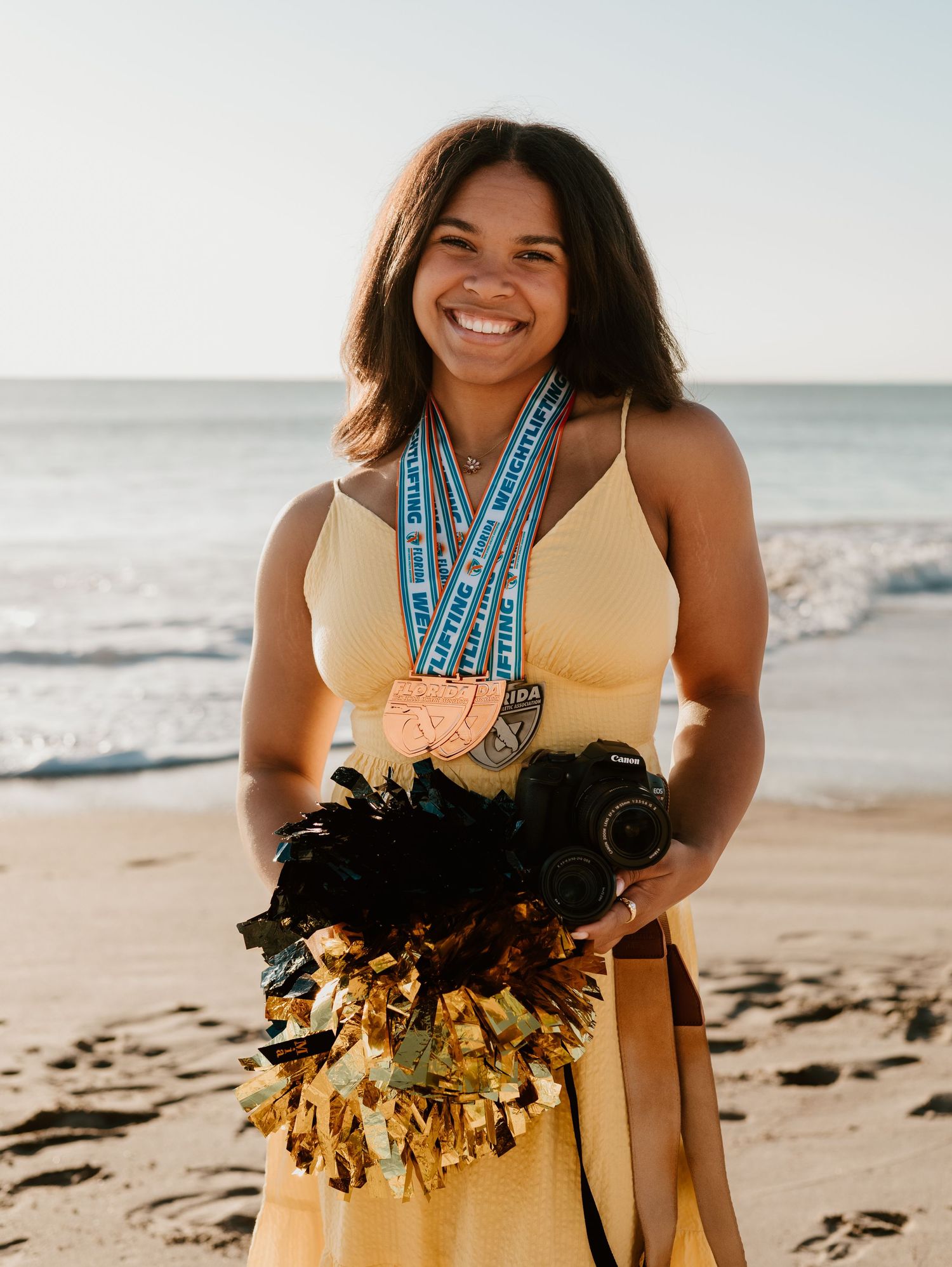 A cheerful person in a yellow dress with medals and a grass skirt poses on a sunny beach.