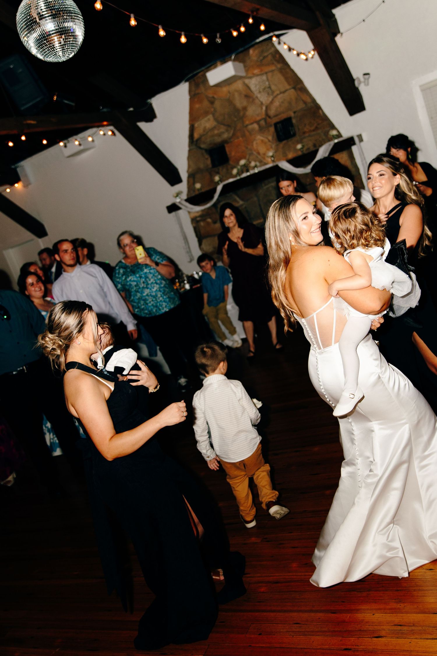 Bride in white gown joyfully holds child on dance floor at lively wedding reception celebration.