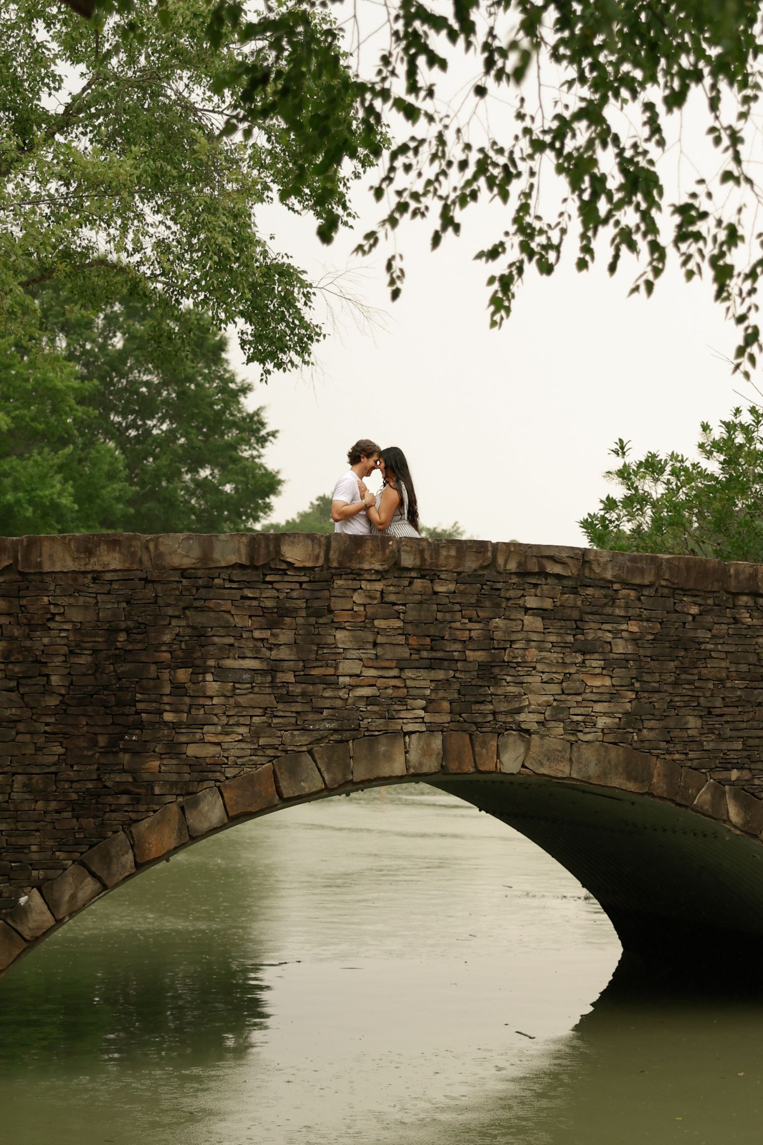 Couple sharing a romantic kiss on a rustic stone arch bridge over a calm river, surrounded by lush green trees.