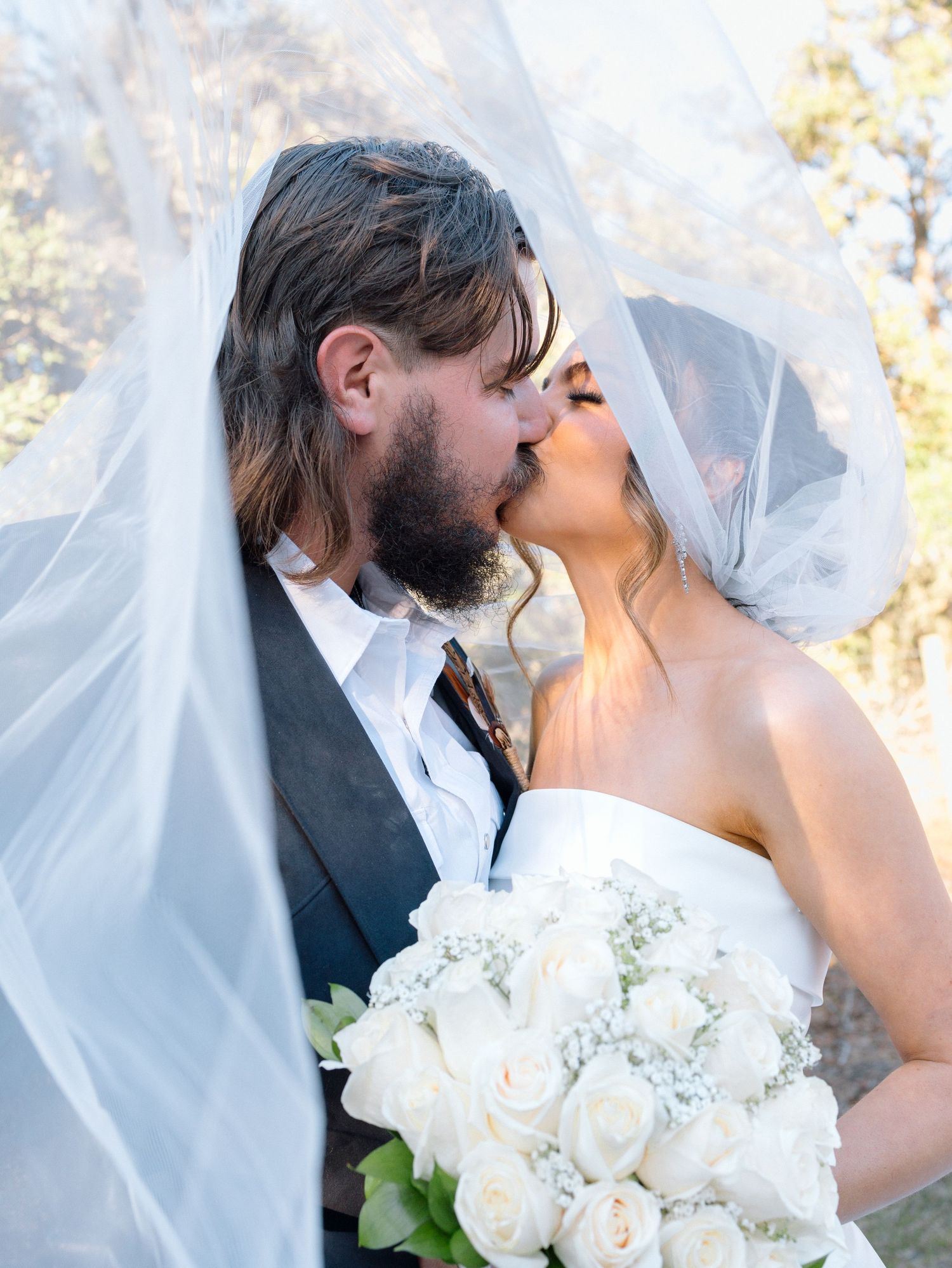 A romantic wedding photo showing a couple kissing under a flowing white veil while holding a bouquet of white roses.