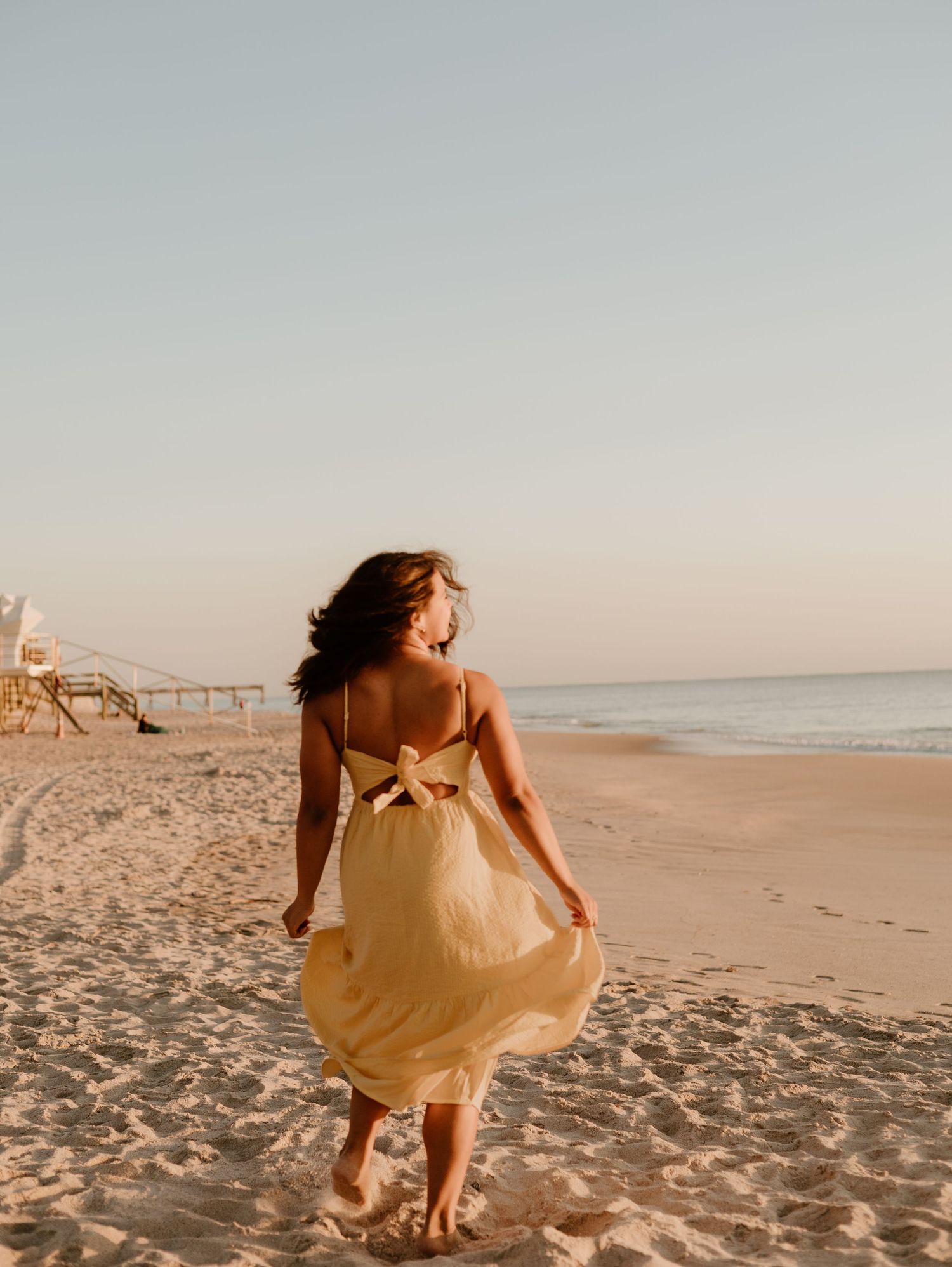Woman in yellow sundress walking barefoot on sandy beach at golden hour with ocean waves in background.