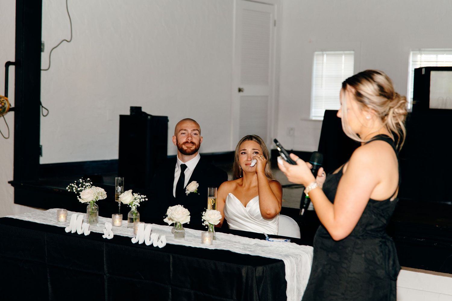 Bride and groom laughing at head table while bridesmaid gives wedding speech at elegant black and white reception.