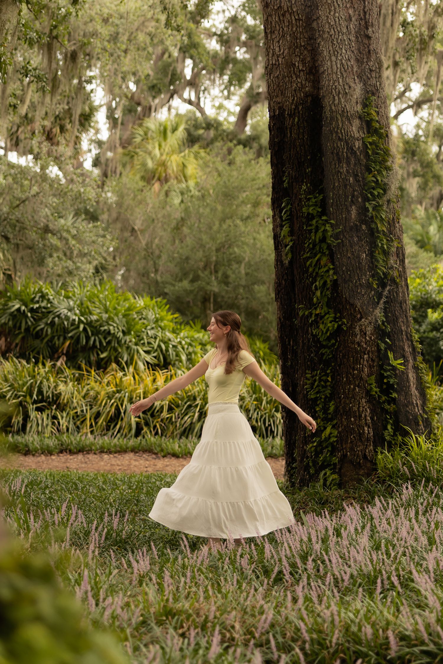 A woman in a white tiered skirt and yellow top twirls gracefully in a lush green garden near a large tree.