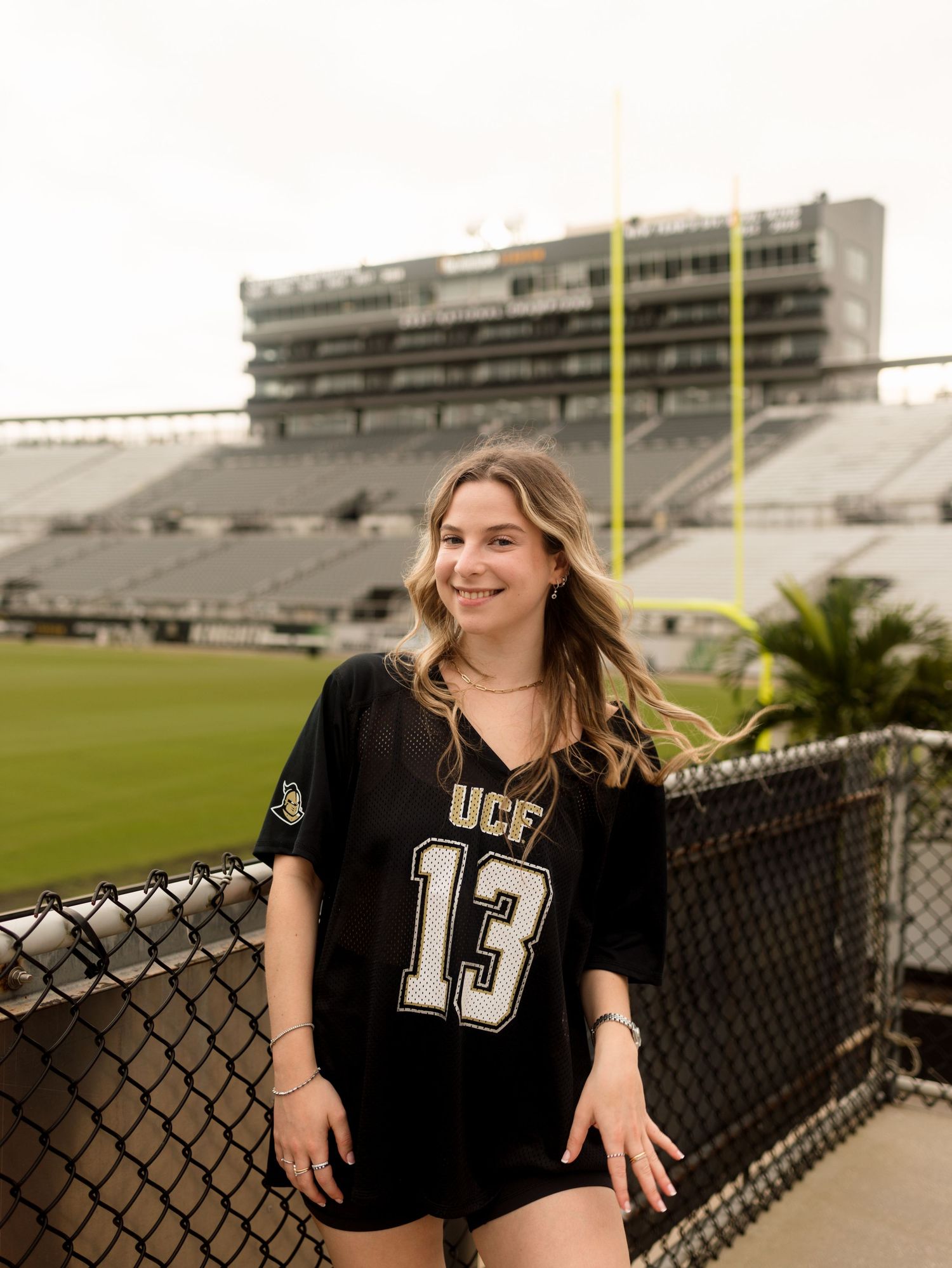 Young woman in UCF #13 jersey smiling at football stadium, standing by chain-link fence with field and stands behind her.