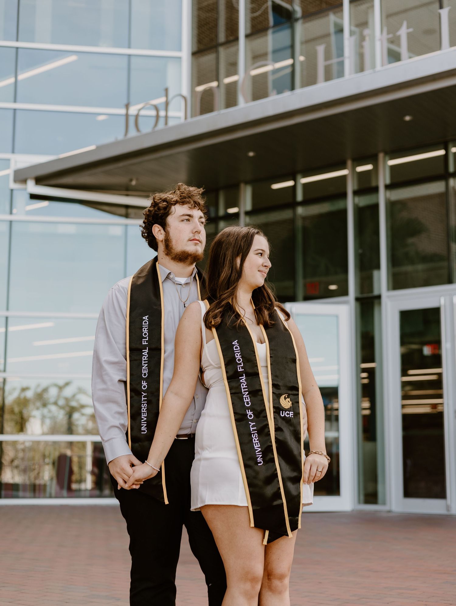 Two UCF graduates in black and gold University of Central Florida stoles pose holding hands outside a campus building.