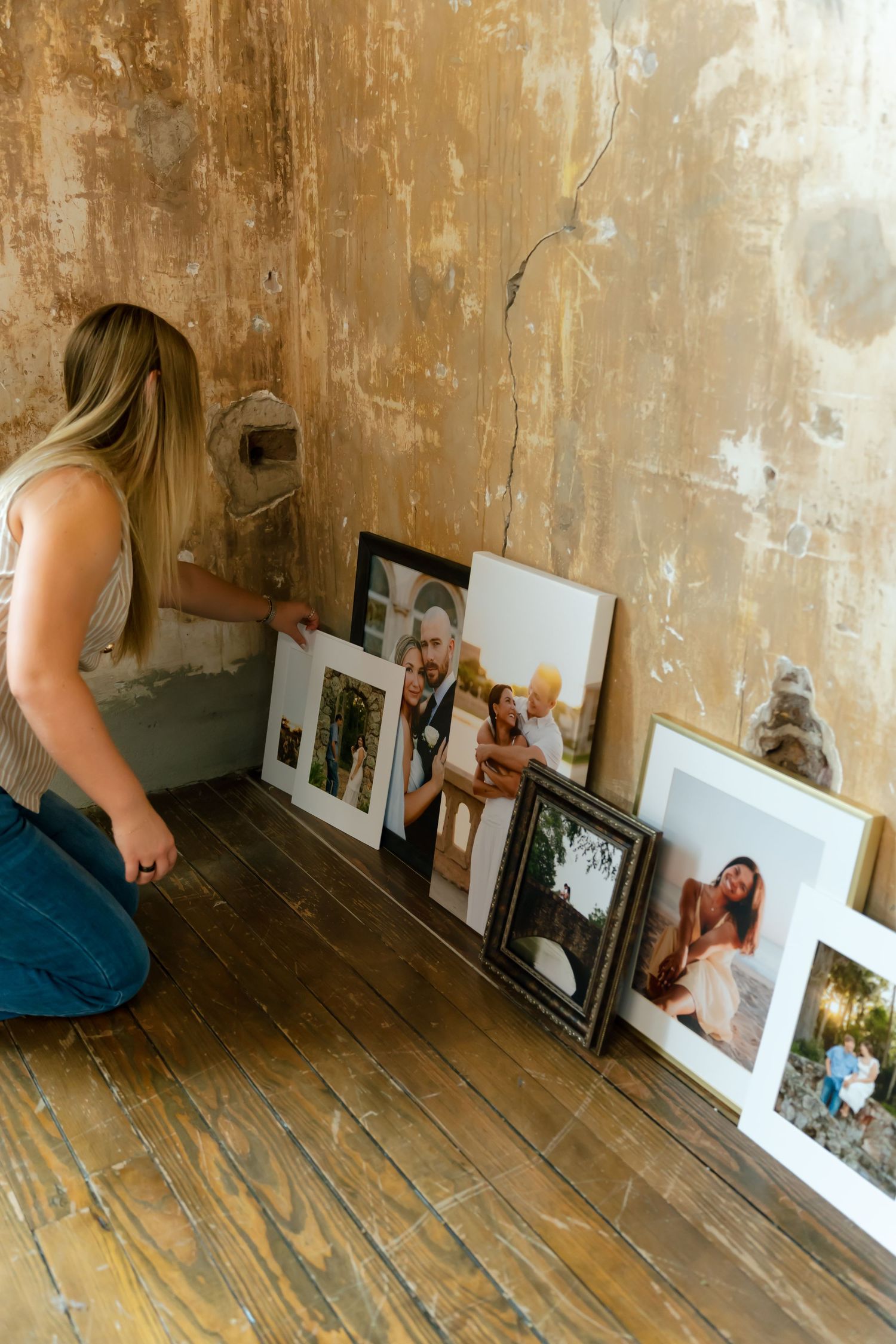 Woman arranging framed family and wedding photos leaning against a rustic distressed wall on a wooden floor.