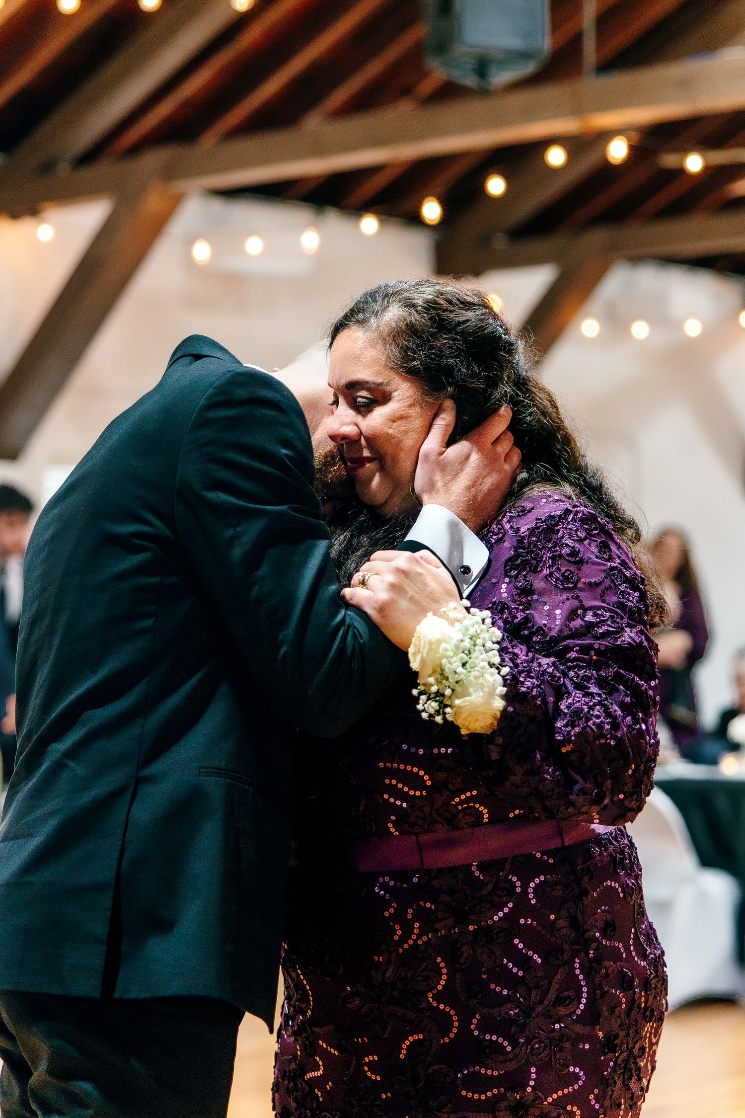 A groom shares an emotional dance with his mother in a purple lace dress at a wedding reception with string lights.