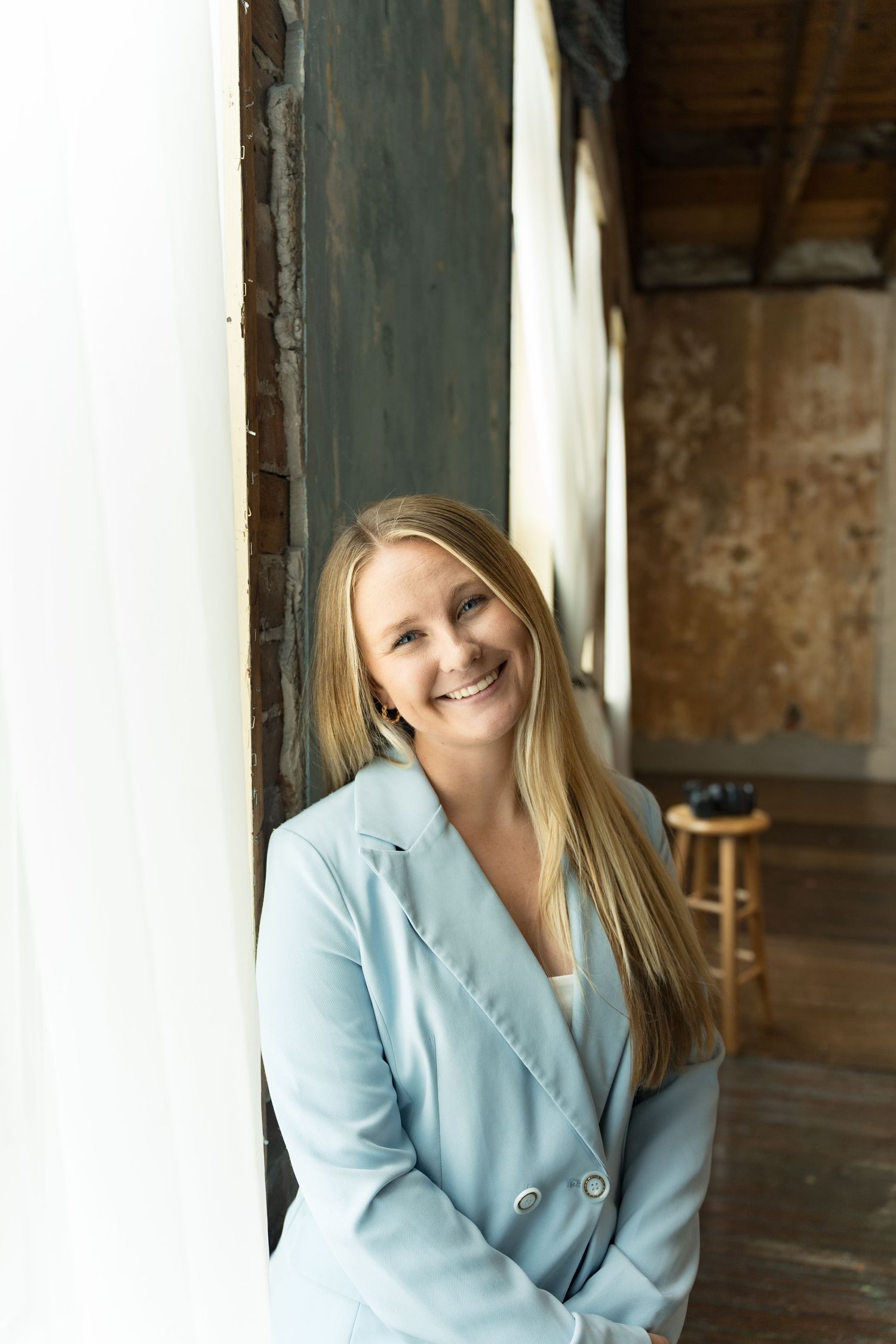 Smiling blonde woman in a light blue blazer leaning against a white pillar in a rustic interior setting.