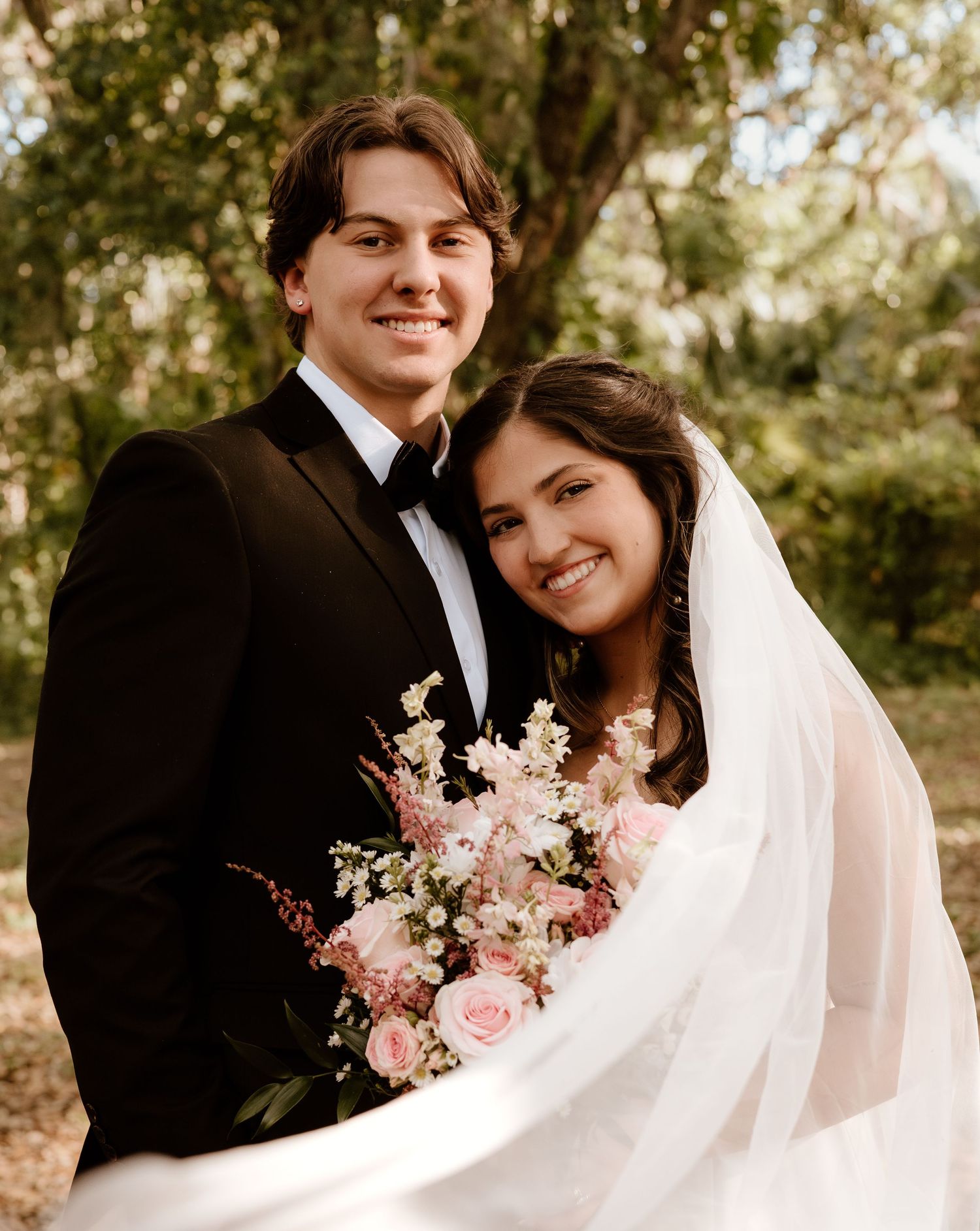 A newlywed couple shares intimate moments in a wooded setting with pink floral accents.