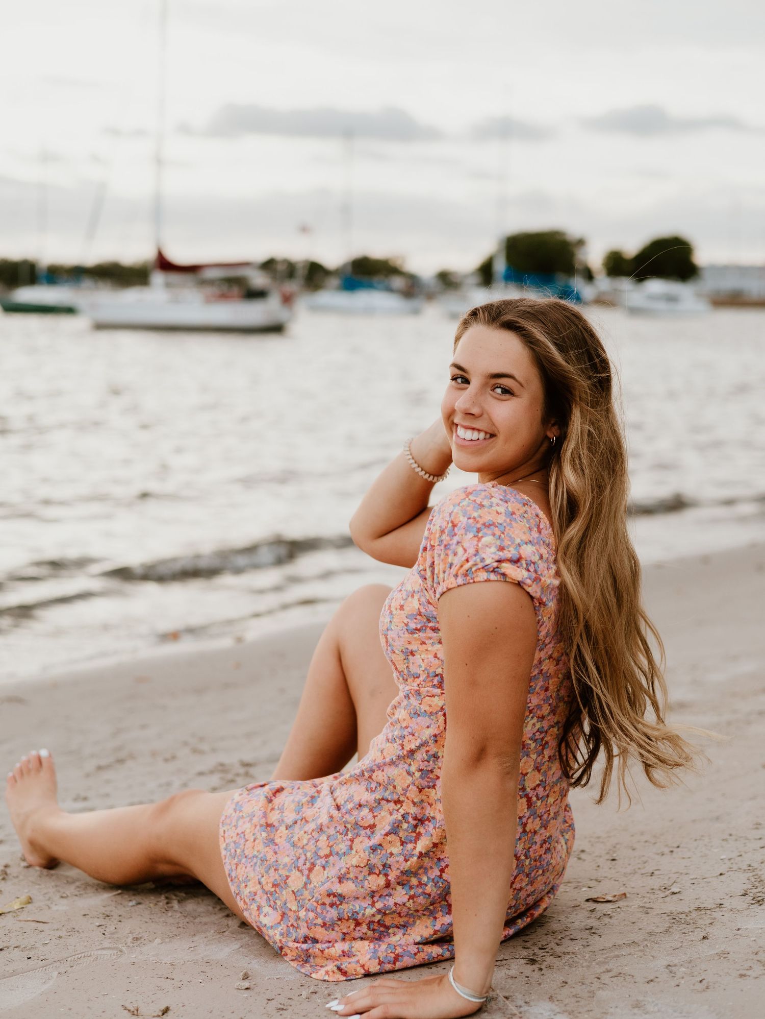 A woman in a floral dress poses on a sandy beach with boats and water in the background.