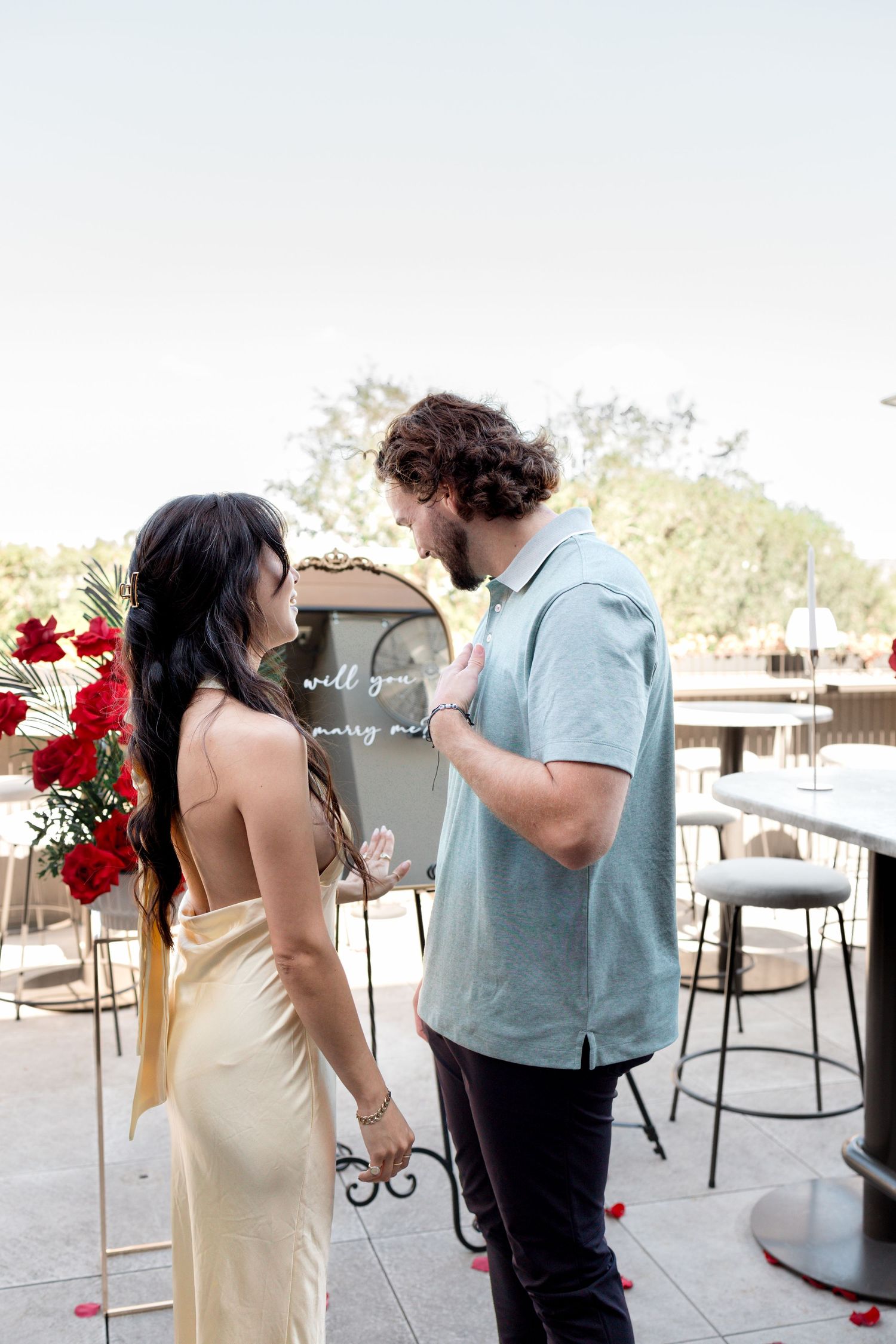 A couple shares a tender moment at an outdoor venue with red floral decorations and bar stools visible in the background.