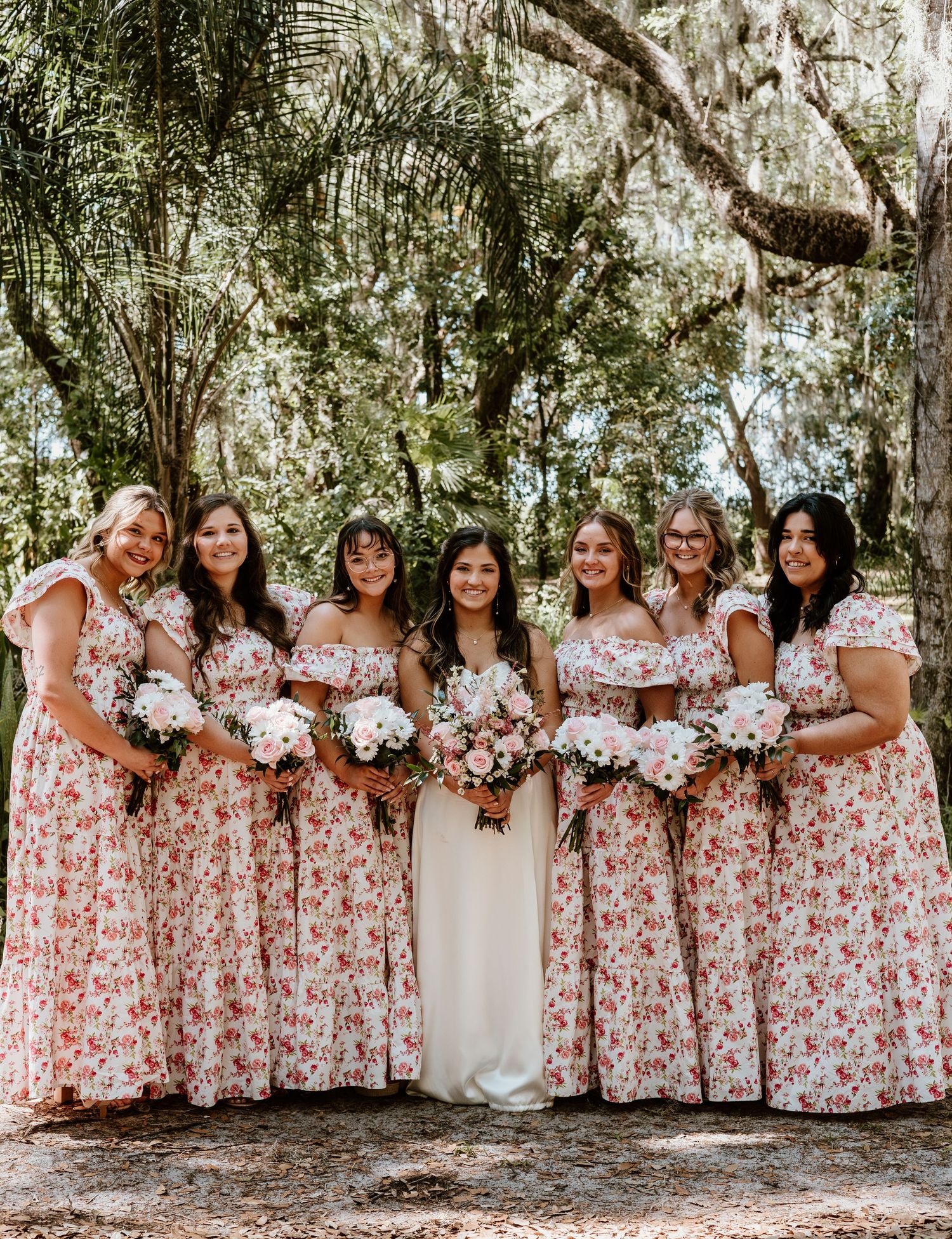 Bridesmaids wearing matching floral pink and white dresses pose together in an outdoor garden setting.