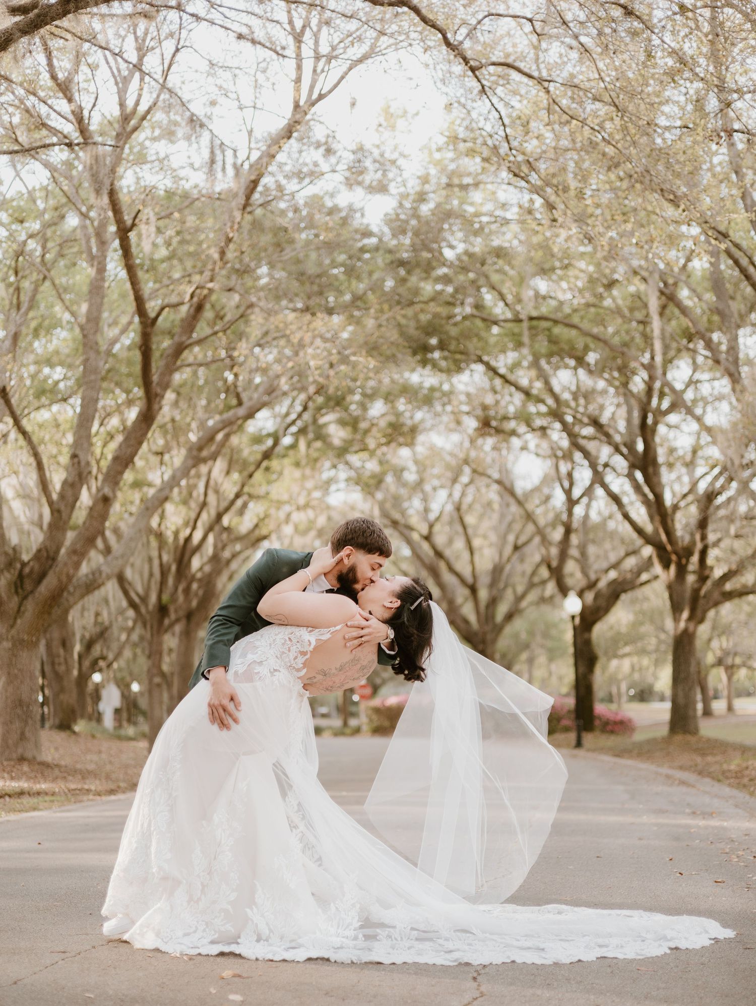 Couple sharing a romantic kiss during their outdoor wedding under oak trees with a flowing white lace dress and veil.