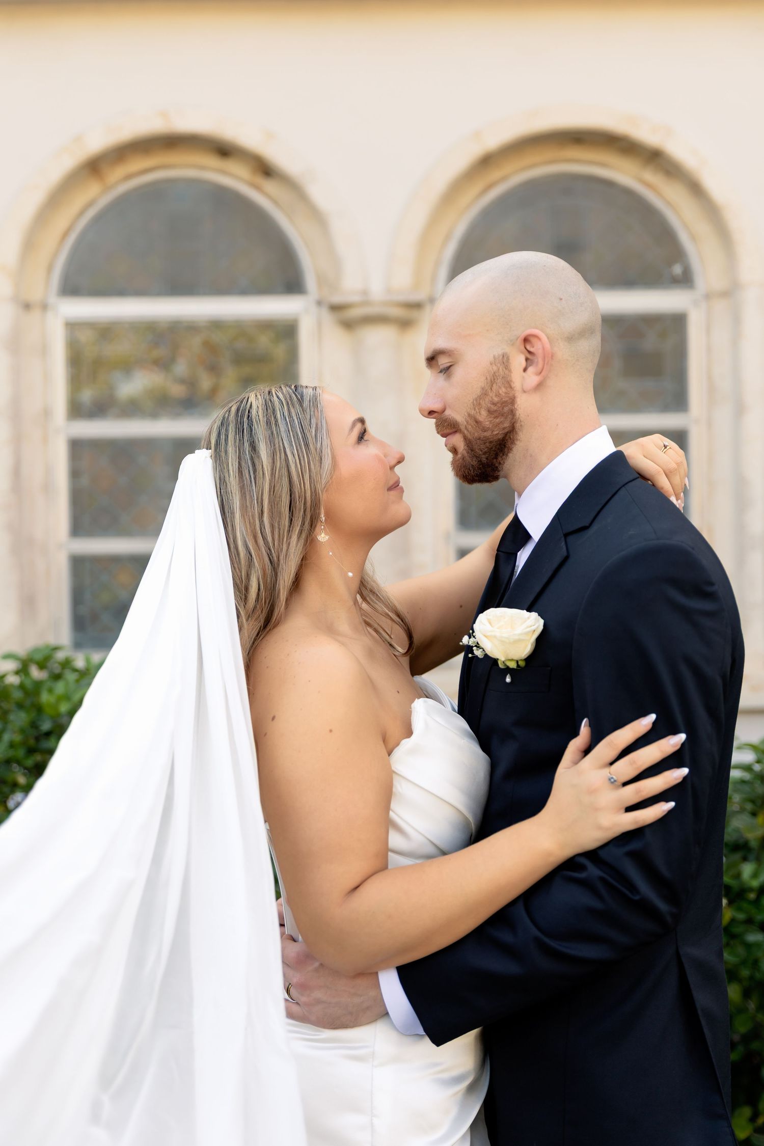 Bride and groom embracing outdoors, gazing into each other's eyes, wearing white gown with veil and black suit with boutonniere.