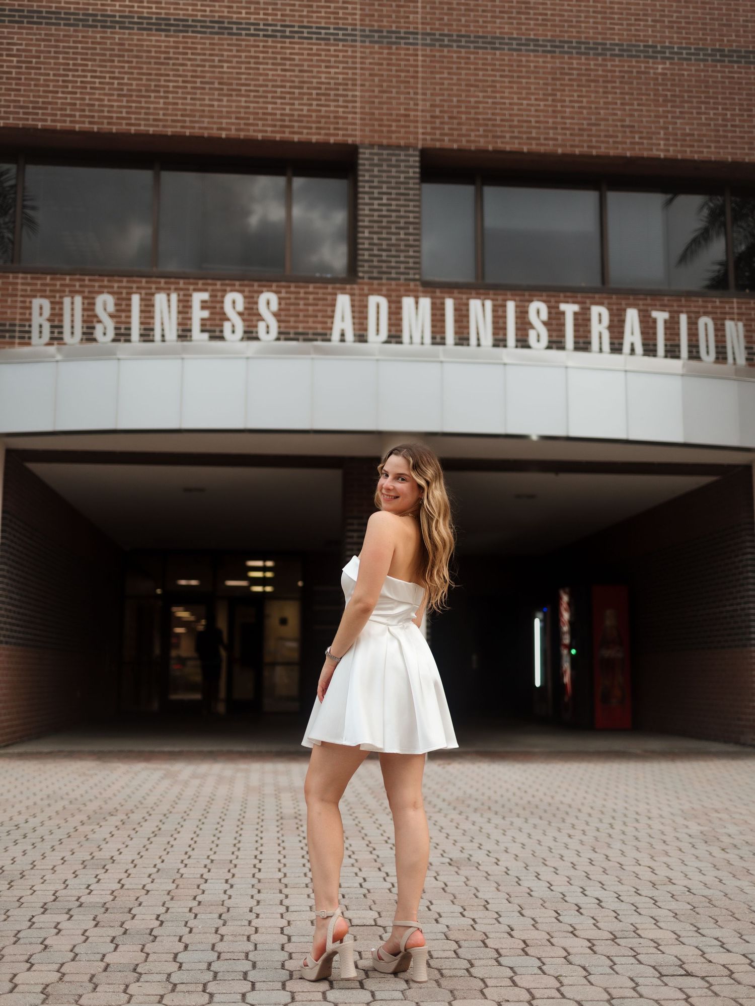 Young woman in white mini dress and heels posing in front of a Business Administration building.