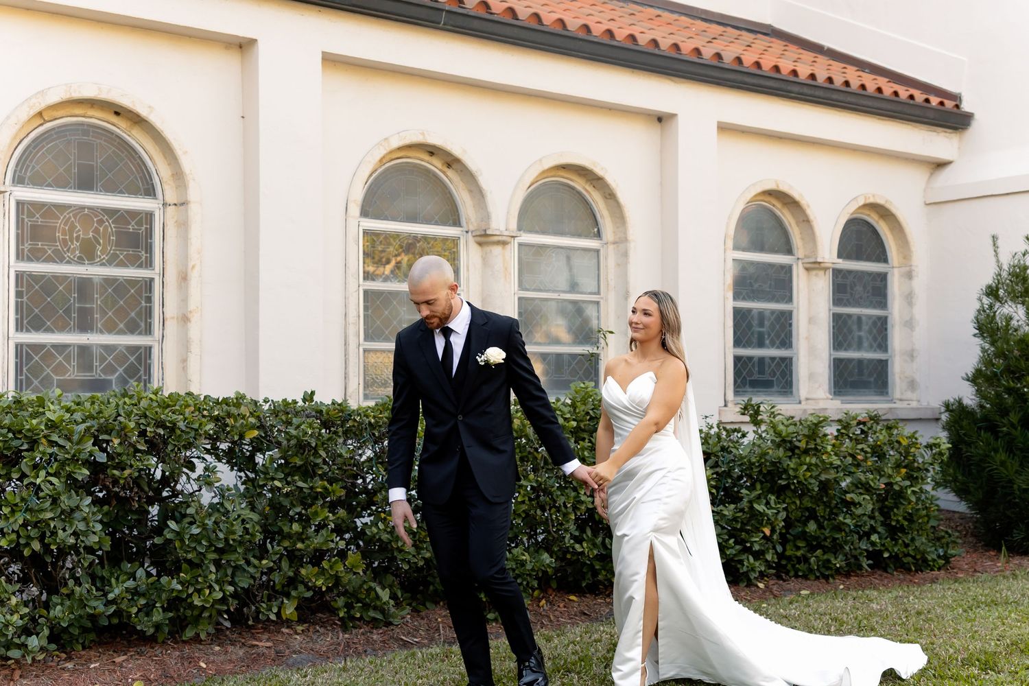 A bride and groom walk hand in hand outside a white building with arched windows on their wedding day.