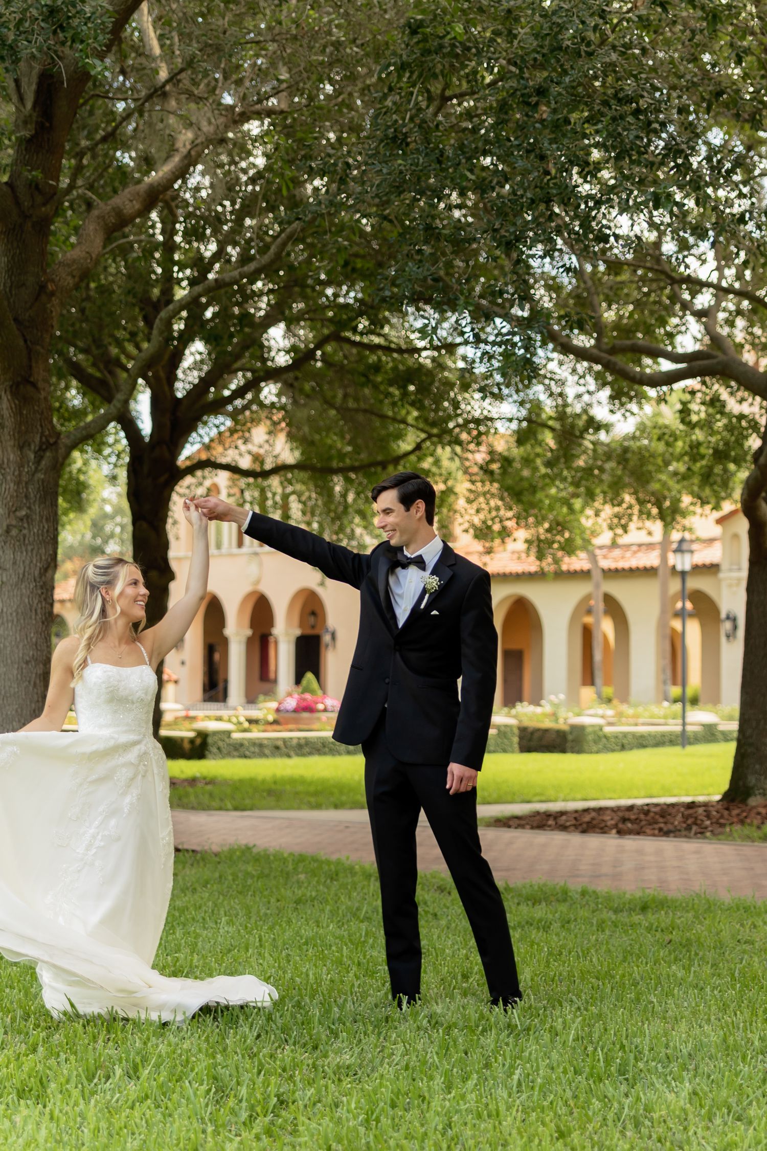 Bride in white gown and groom in black tuxedo dancing on green lawn with arched building backdrop.
