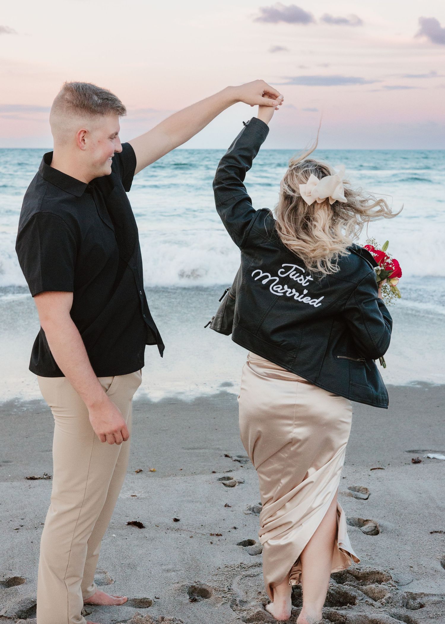 Couple in matching black shirts dancing playfully on a beach at sunset.