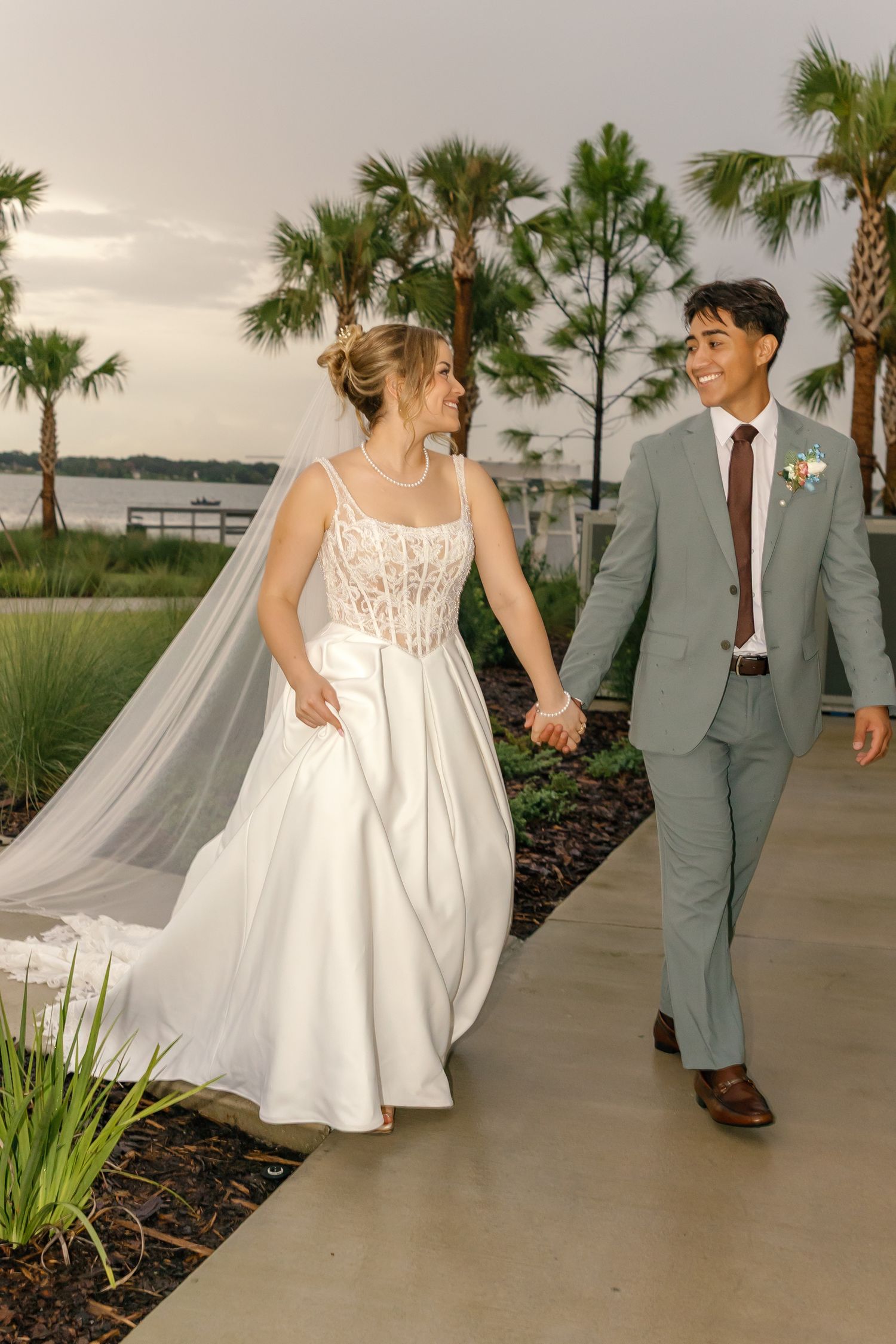 A bride and groom walk hand-in-hand outdoors near palm trees and waterfront, smiling at each other on their wedding day.
