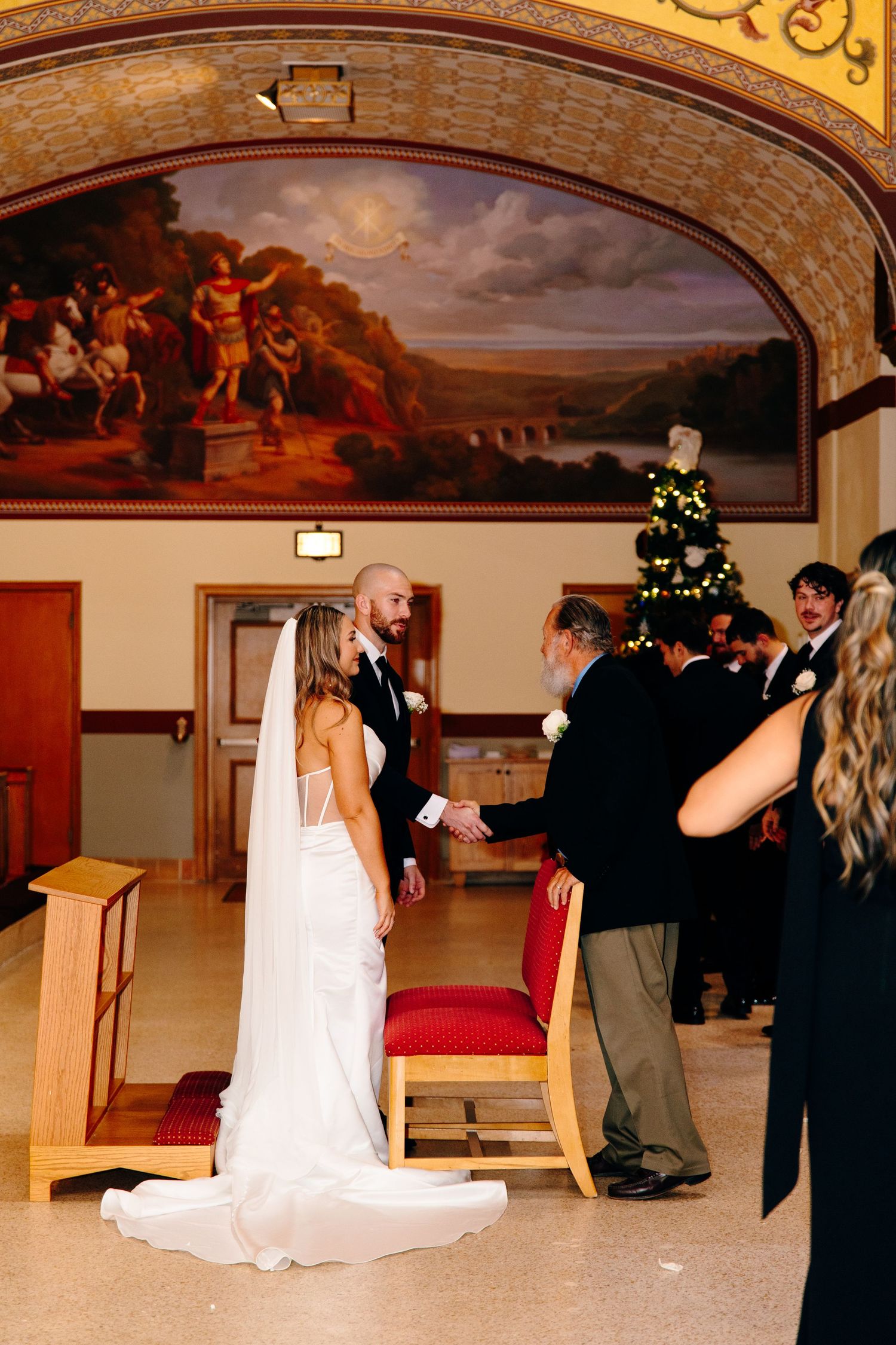 Bride and groom exchange vows at elegant wedding ceremony beneath ornate arch with classical mural.